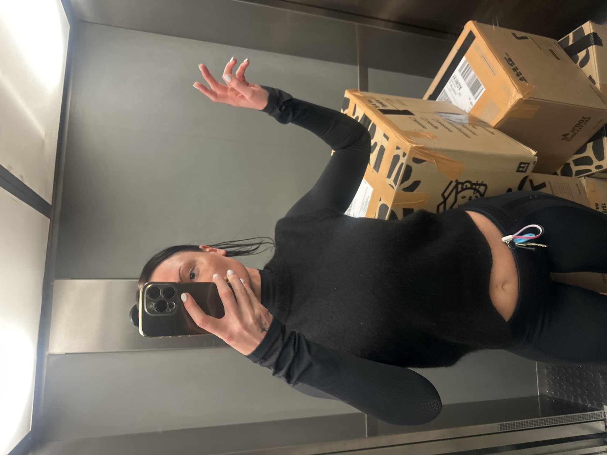 An individual, appearing female, is depicted in a selfie taken within an enclosed space, most likely an elevator, characterized by smooth, grey metallic wall panels and overhead fluorescent lighting. She wears a black long-sleeved, high-neck cropped top and black athletic pants, exposing her midriff. Her dark hair is pulled back. She holds a dark-colored mobile phone, identified as an iPhone Pro model with a gold-colored frame and multiple rear cameras, in her right hand, partially obscuring her face. Her left hand is raised and splayed, displaying white nail polish, and a small star-shaped tattoo is visible on her left wrist. A set of keys with blue and pink fobs hangs from her left hip. To her right, a stack of multiple brown cardboard boxes is present. One box features a black and white abstract or patterned design, while others display orange sealing tape and visible branding including "DHL" and "ADIDAS Originals." The precise location in London, United Kingdom, cannot be determined from the visual information provided.