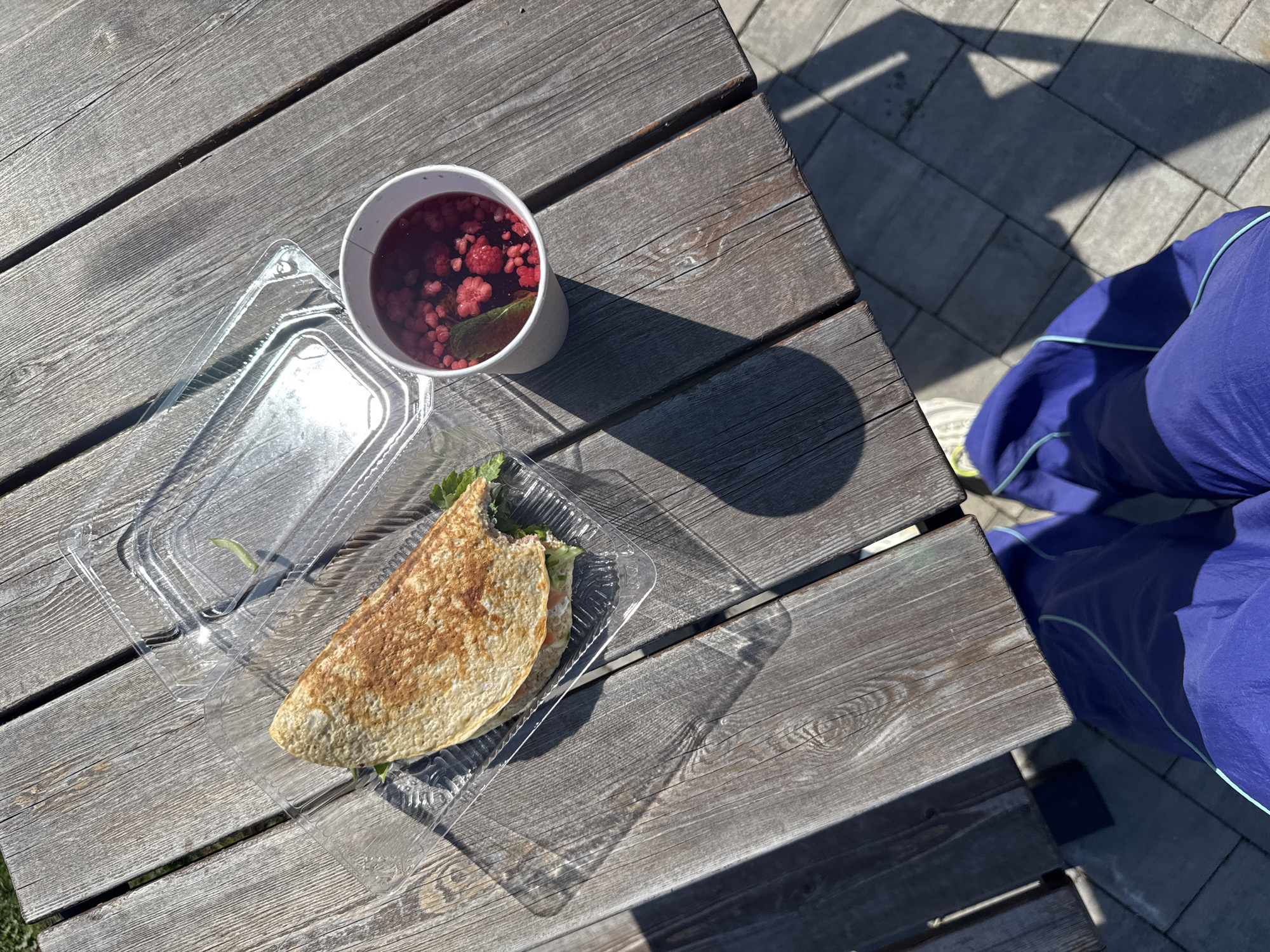 The image shows a wooden picnic table outdoors with a meal of a crepe in a plastic container and a cup of berry drink. There are a lot of shadows, indicating it is sunny. The location may be in the city of Krasnoyarsk, Russia. There is a person wearing blue pants with a turquoise stripe sitting at the table.