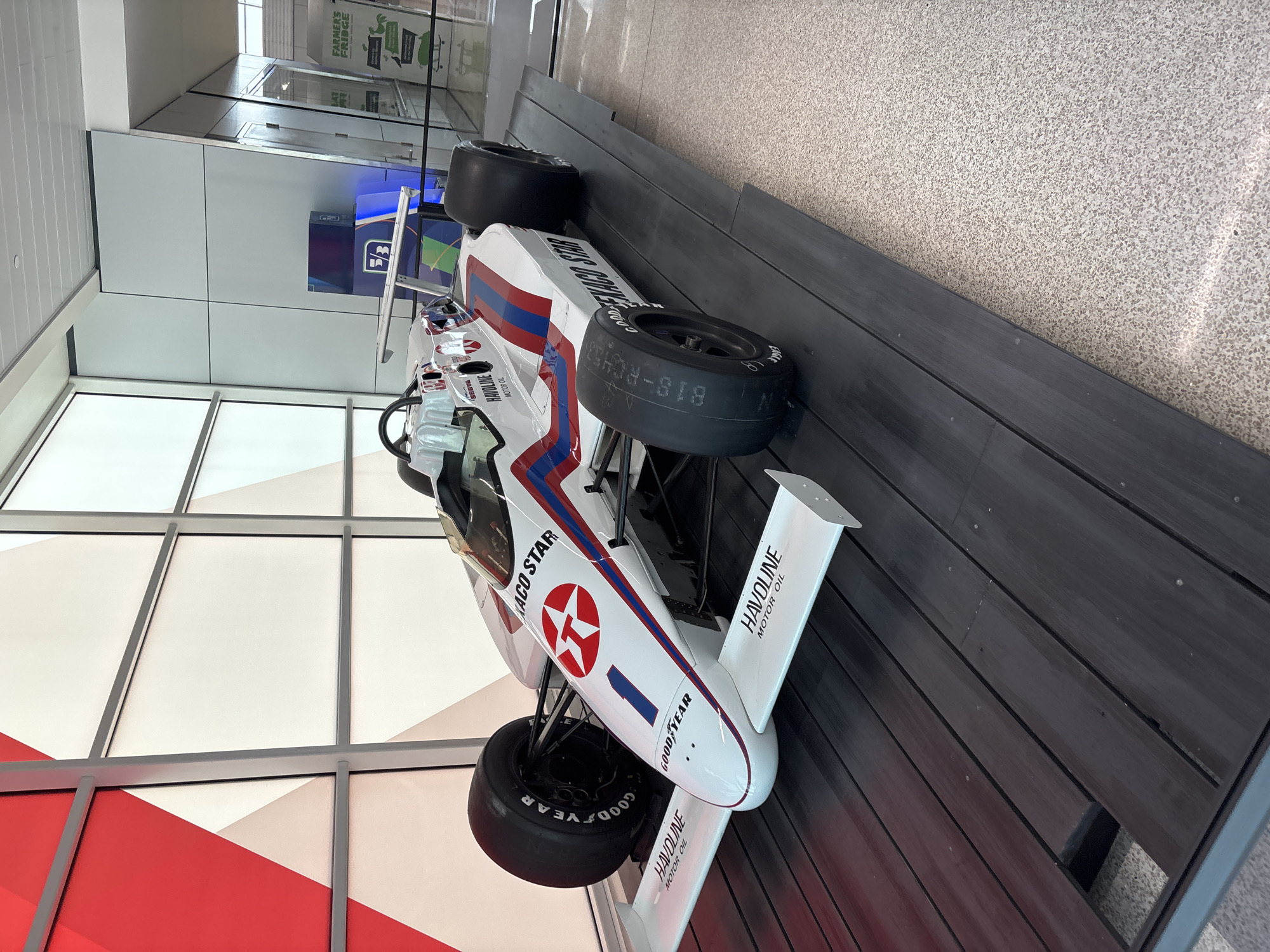 The image shows a vintage race car on display. The car is white with red and blue stripes, and it has the logos of Texaco Star and Havoline on it. The tires are Goodyear brand. The car is sitting on a dark wood platform. In the background, there is a large window with white and red panels, and a sign for "Farmer's Fridge". The location is likely in the city of Indianapolis, United States of America.