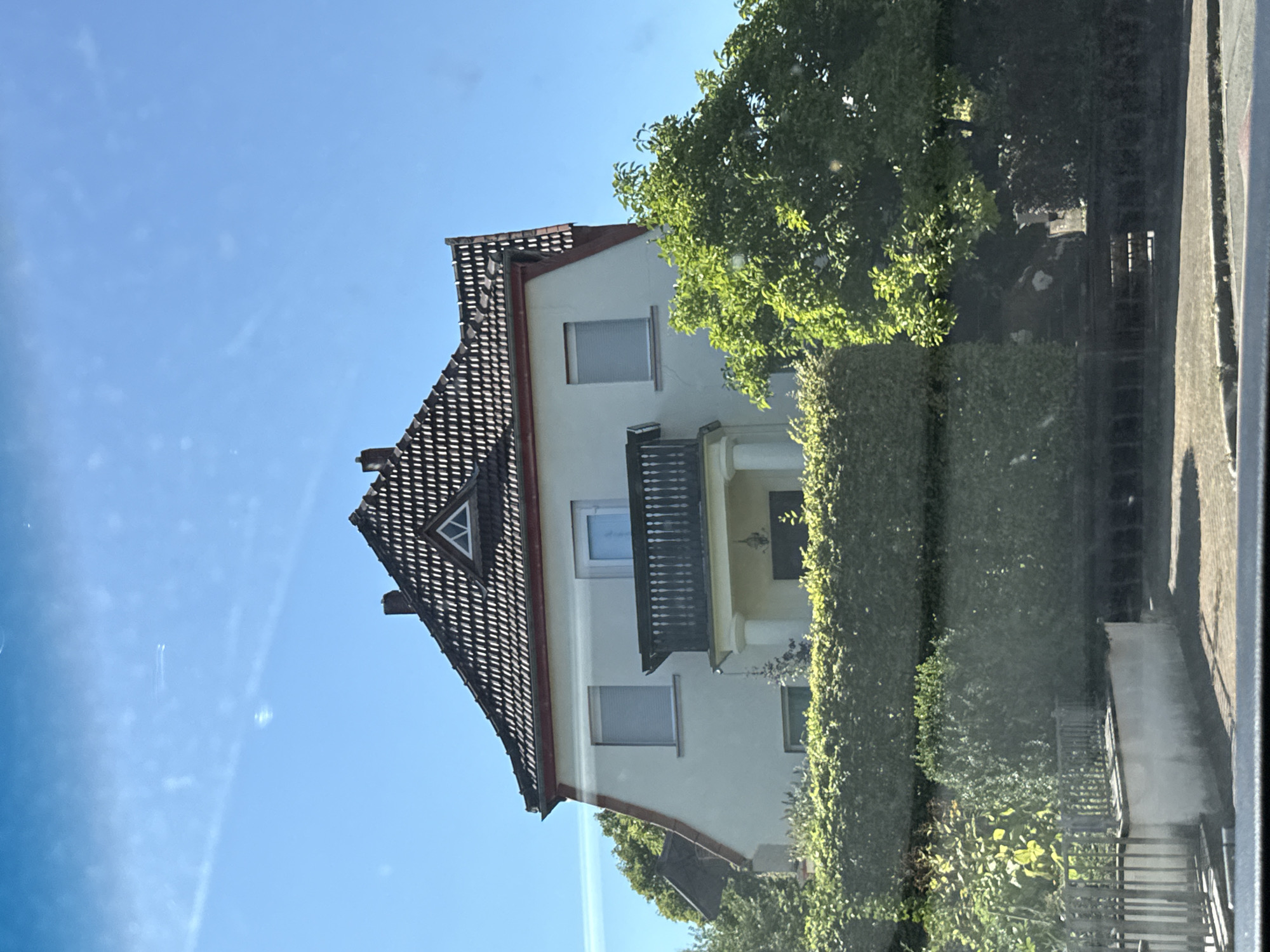 An exterior, eye-level, medium shot captures a detached, multi-story house situated in a residential area, likely in the town of Bad Sachsa, Germany. The house has a light-colored facade, a dark-tiled, steeply pitched roof with a prominent dormer window, and a red trim along the roofline. A balcony with a dark railing and pillars is visible on the upper floor, above the entrance. Lush green trees and manicured hedges surround the property, with a low metal fence and a concrete wall in the foreground. The sky is clear blue, with some faint contrails visible. The lighting suggests a bright, sunny day.