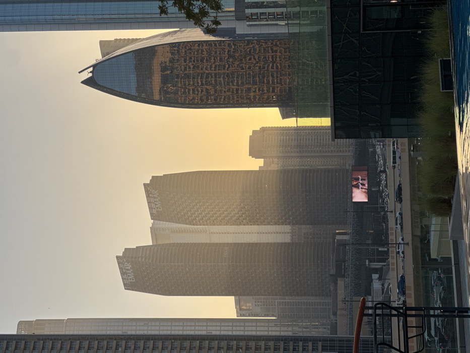 The image displays an urban cityscape in Dubai, United Arab Emirates, during either sunrise or sunset, indicated by a warm, hazy, golden-yellow sky. In the immediate foreground, the edge of a swimming pool is visible on the lower right, reflecting the sky. To the lower left, a dark metal railing partially obstructs the view of a paved area with green foliage in planters. The midground features an elevated multi-lane highway with moderate vehicle traffic, including cars, vans, and buses, moving in both directions. Below the elevated highway, additional vehicles are parked. A large digital billboard, positioned above the highway, displays an advertisement featuring a person, likely a woman, with Arabic text. The background is dominated by numerous high-rise buildings. Two prominent, dark skyscrapers with textured facades, bearing the "EMAAR" logo near their tops, are situated left of center. To the right, a distinctive dark glass skyscraper with a curved, pointed apex reflects distorted images of other buildings and the sky. A tall, slender building is partially visible on the far left. No individuals are discernible in the foreground or midground.