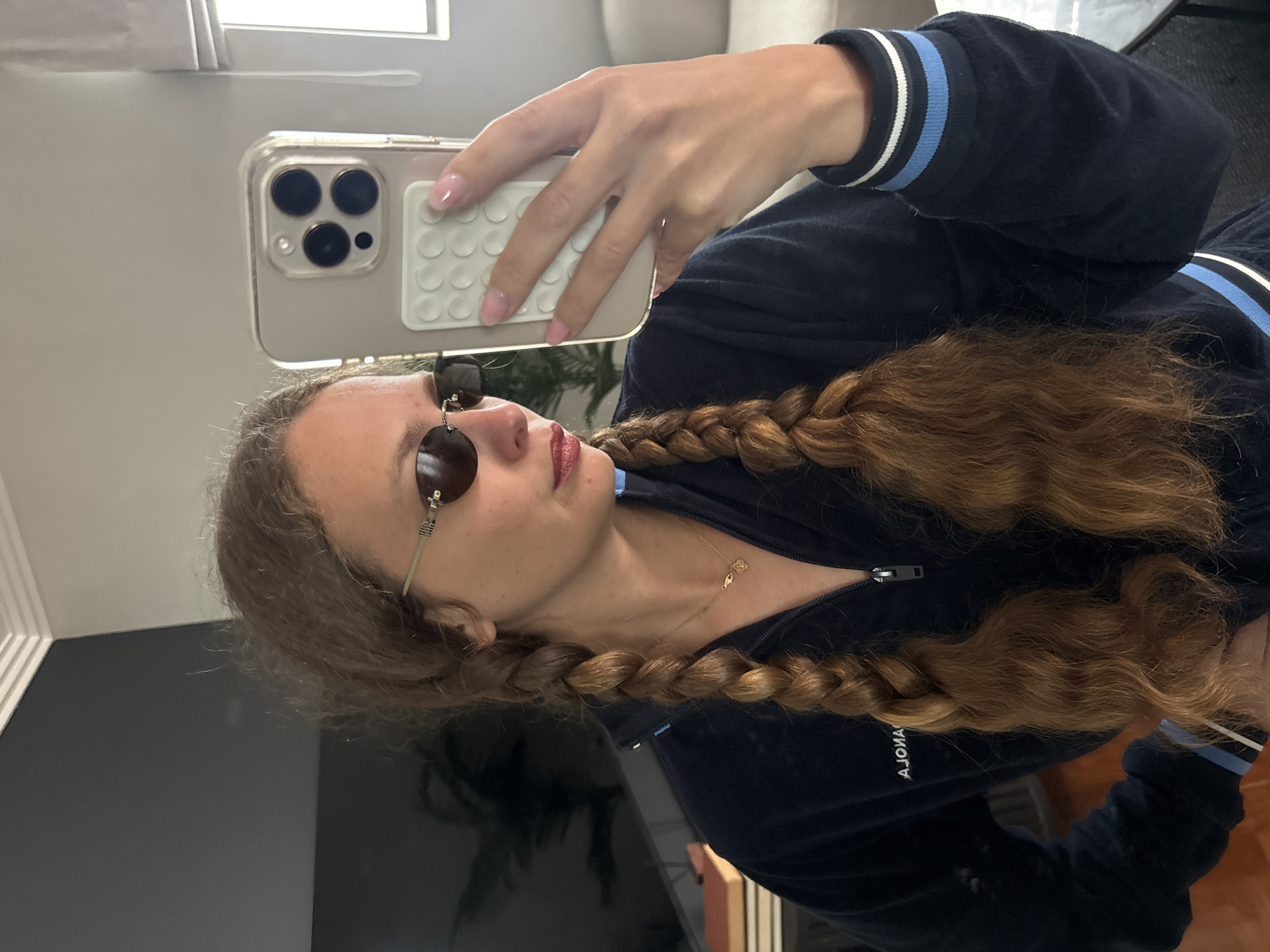 A young woman is taking a selfie. She has light brown hair styled in two braids and is wearing sunglasses with round lenses. She is wearing a navy blue velour tracksuit with blue and white striped trim on the sleeves. She is holding a smartphone in her right hand to take the photo. The phone is in a clear case with a white accessory on the back. The background is an interior scene with a window providing natural light. There is furniture visible, but the specific details are not clear. The city location is not discernible from the image.