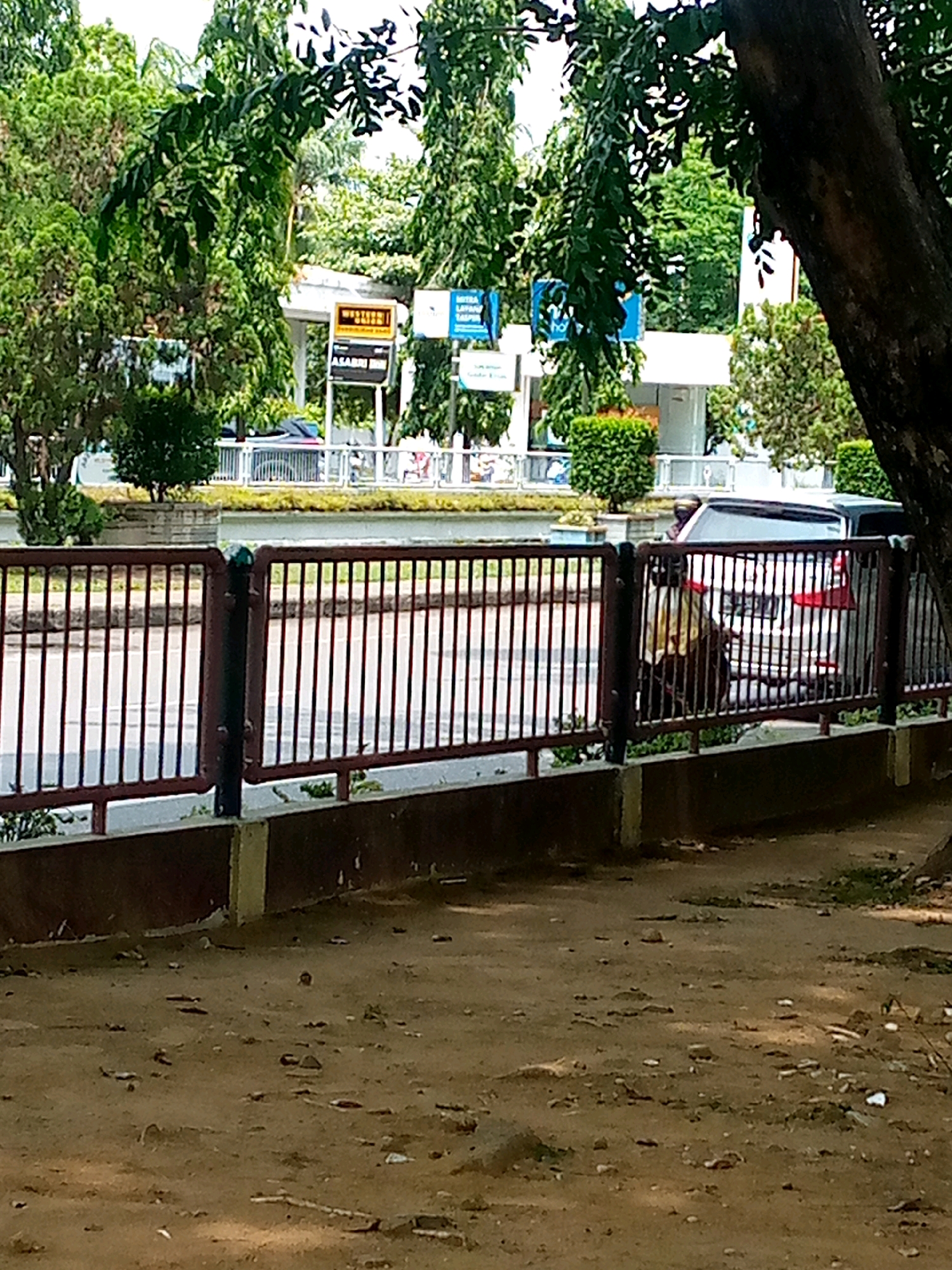 The image shows a street scene in Lhokseumawe, Indonesia, with a focus on a metal fence bordering a sidewalk and a road. The fence is painted a dark red and is supported by dark vertical posts. Beyond the fence, a road is visible with a dark-colored vehicle, possibly an SUV, parked on the right side. A person wearing a yellow shirt and a dark helmet is visible standing next to the vehicle. Further down the road, there are signs, one of which appears to be a "Western Union" sign, suggesting a commercial area. The background is filled with lush green trees and foliage, indicating a tropical environment. The foreground consists of a sandy, dirt ground with scattered debris. The overall impression is a glimpse of everyday life in an urban area of Lhokseumawe, with a mix of natural and man-made elements.
