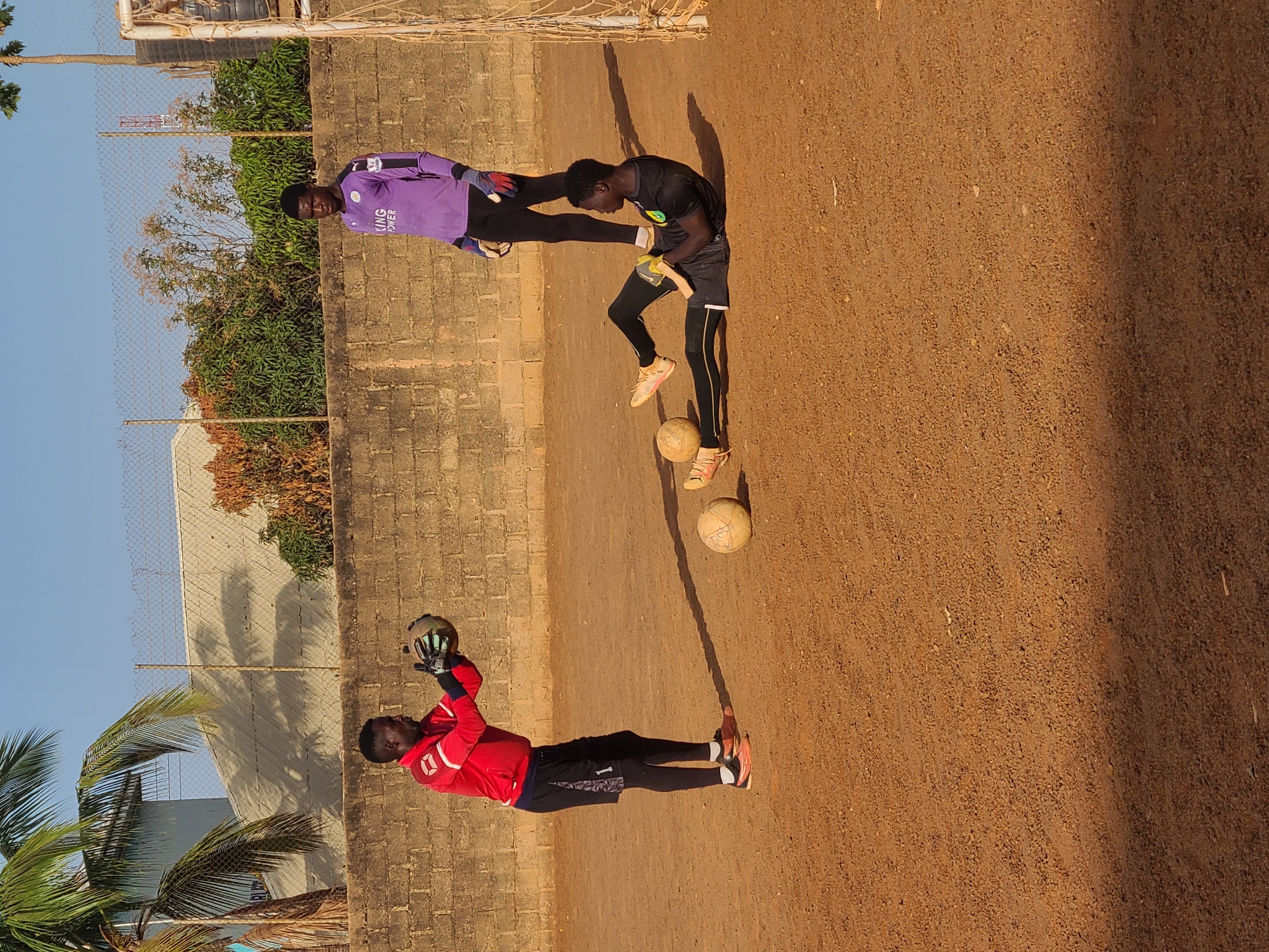Three people are on a dirt playing field in Jos, Nigeria, based on the image's details. The field is bordered by a low brick wall, a chain-link fence, trees, and blue sky. One person is wearing a red soccer jersey with black shorts and catching a soccer ball with gloved hands. Another person is juggling a soccer ball, wearing black athletic leggings and a black shirt. The third person is wearing a purple soccer jersey, black pants, and is standing on the other side of the juggling person. The field is brown dirt, and the lighting indicates it's daytime. A goal net is visible in the upper left corner.