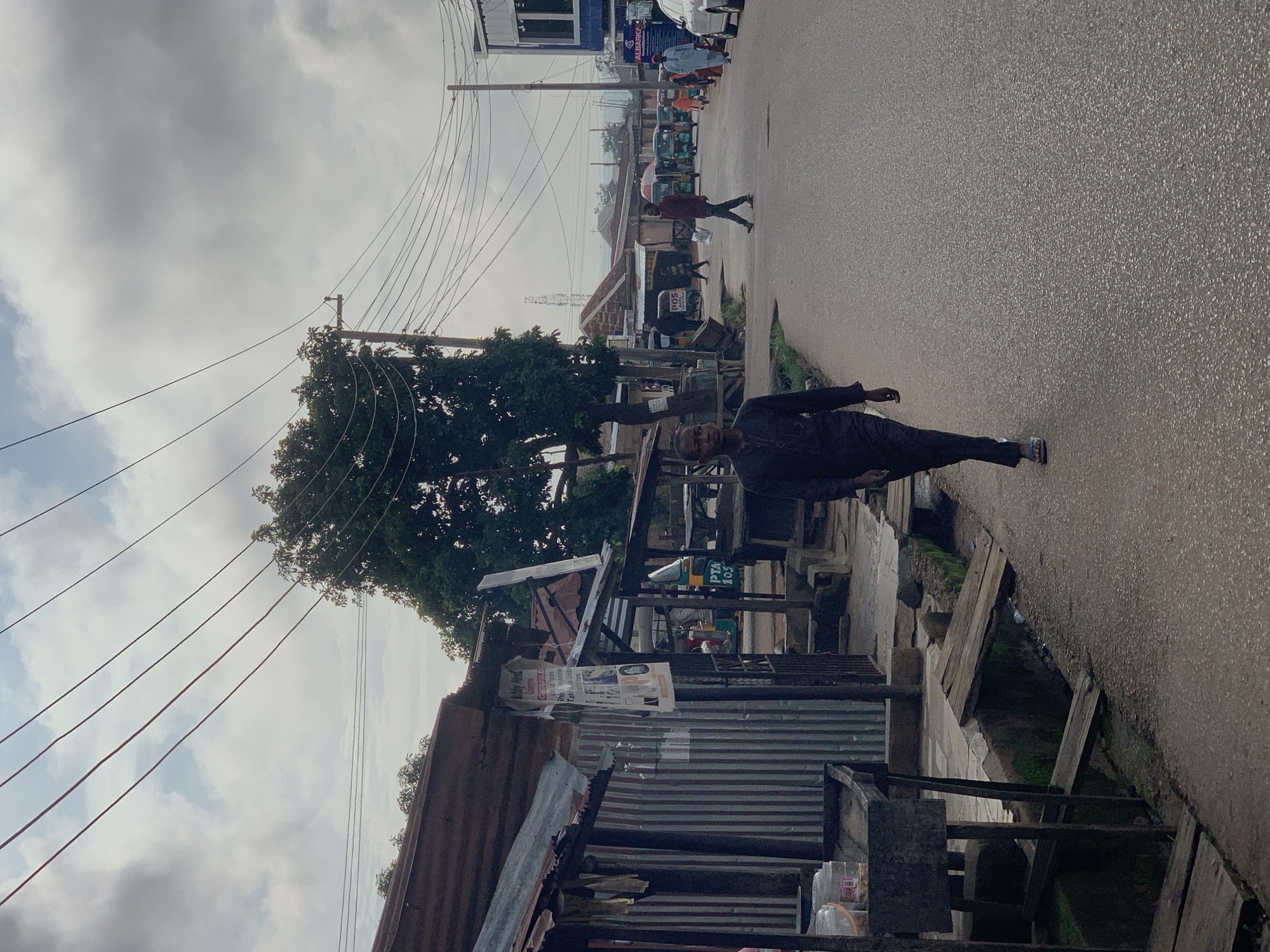 The image captures a street scene in Jos, Nigeria, under an overcast sky. A man in dark clothing walks towards the viewer on an asphalt road. To the left of the road, a row of makeshift structures with corrugated metal roofs and walls is visible, with some wooden planks forming a rudimentary drainage system in front. Various items, including buckets and what appear to be containers, are placed on a wooden stand in front of one of the structures. In the background, several "Keke Napep" (three-wheeled vehicles) are parked or in transit, along with other pedestrians and buildings. Power lines crisscross the sky above the scene. The overall impression is of a busy, informal marketplace or residential area.