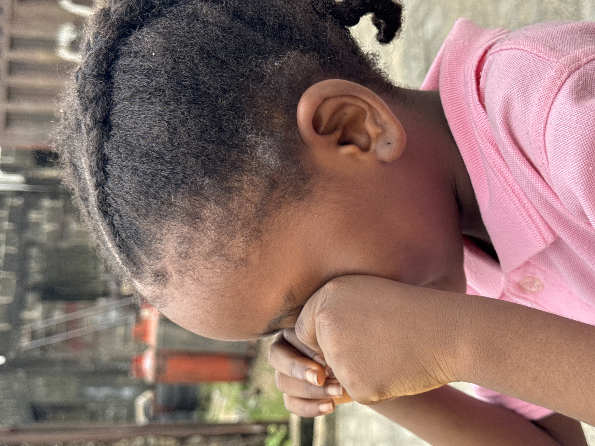 A close-up shot shows a young Black child, likely a girl, with dark, braided hair partially visible. The child is wearing a pink collared shirt. Her head is tilted down and resting on her hand, obscuring her face. The setting seems to be outdoors, in Ado, Nigeria with blurred industrial objects visible in the background.