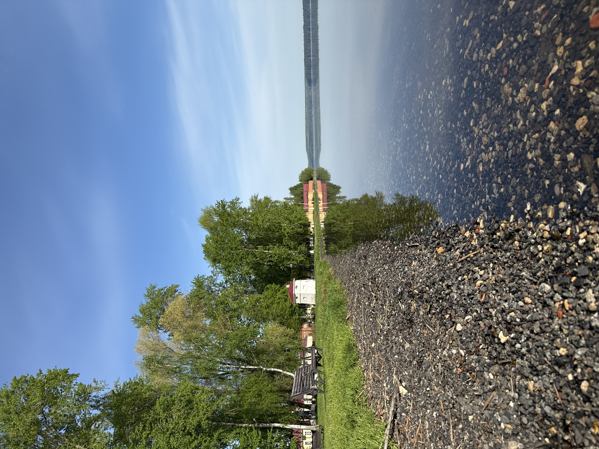 The image depicts a scene at the Votkinsk Reservoir in Votkinsk, Russia. The perspective is a shoreline view, with the reservoir visible on the right half of the frame, and the land on the left.
The reservoir appears calm, with reflections of the sky and distant land masses visible. The water's edge transitions into a rocky shoreline with gravel and small stones.
On the land side, there's a grassy area leading to trees, benches, and a small building with a red roof. The sky is blue with some light clouds. There are no people visible in the frame.