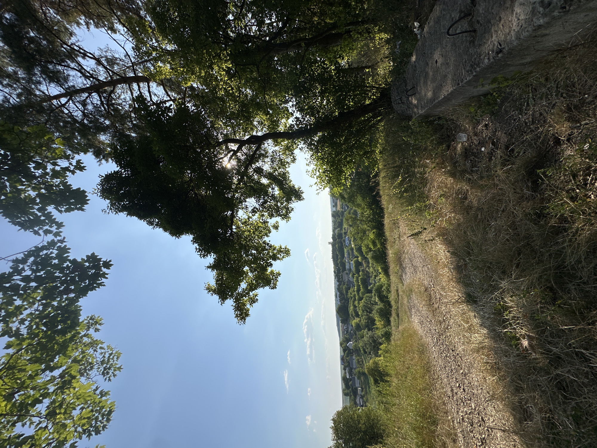 A sun-drenched view of a rural landscape is captured from an elevated position. In the foreground, a gravel path leads down a grassy slope, flanked by dry grass and shadows cast by trees. To the right, a weathered concrete structure with exposed rebar juts out into the frame. Beyond the immediate foreground, the terrain slopes down to reveal a village nestled amongst lush green trees. White-roofed houses are scattered throughout the valley, with hints of a church steeple visible in the distance. The sky above is a clear, bright blue with a few scattered white clouds. The overall impression is one of a serene, sunlit day in the Ukrainian countryside, likely in or near Makiv.