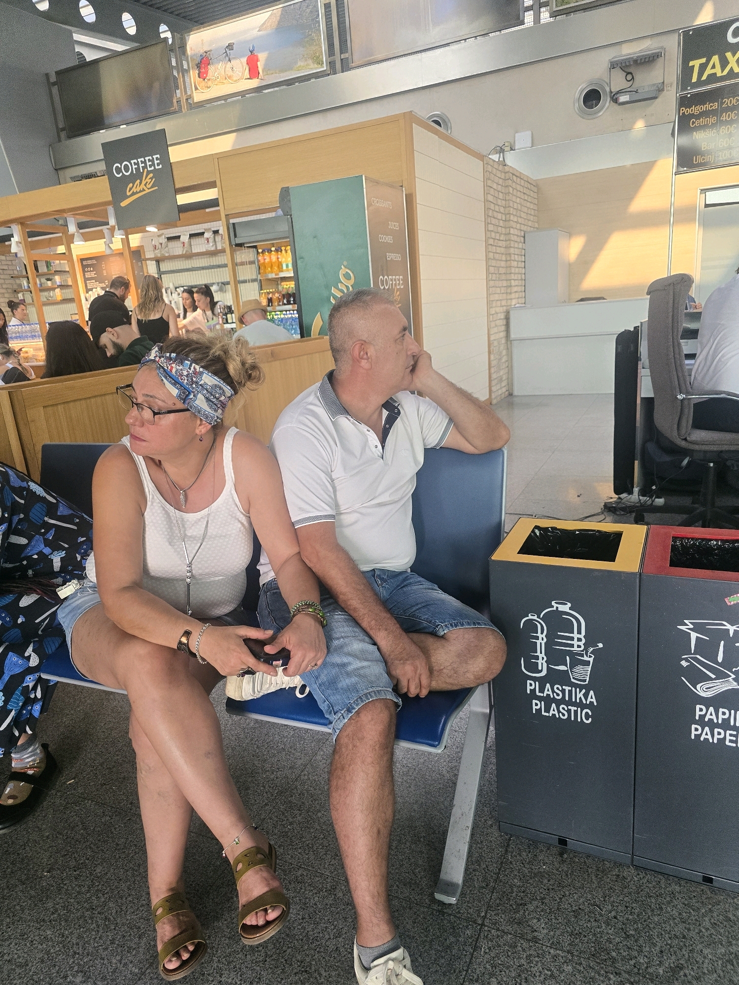 The image depicts a man and a woman sitting on a bench in what appears to be an airport or bus station waiting area. The woman is on the left, wearing a white tank top, denim shorts, glasses, and a patterned headband. She has jewelry on her wrists and neck. The man is to her right, wearing a white polo shirt and denim shorts. He is resting his head on his hand, looking to the side. In the background, there is a coffee shop with a sign that reads "COFFEE cake". There are also recycling bins labeled "PLASTIKA PLASTIC" and "PAPIR PAPER" visible. A sign with "TAX" and prices is also visible in the background, which could indicate taxi services available at the location. The overall environment appears to be indoors with overhead lighting. The image may have been taken in Podgorica, Montenegro, as suggested by the presence of a taxi price list that includes destinations within Montenegro, such as Cetinje, Nikšić, Bar, and Ulcinj.