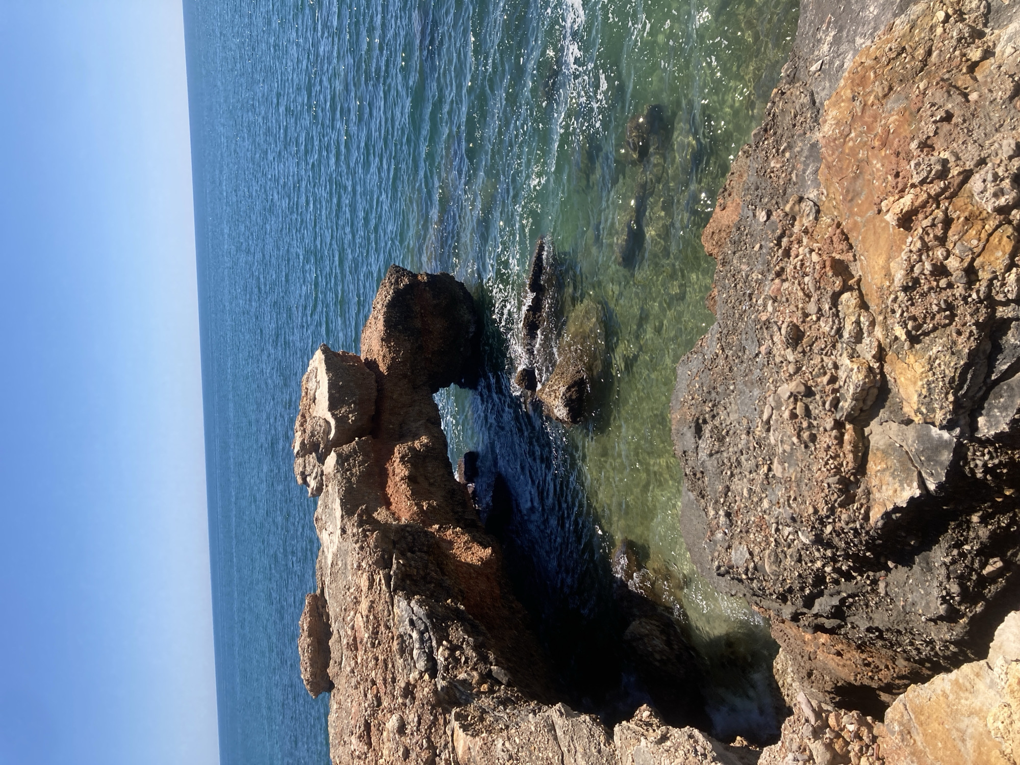 The image shows a rocky coastline with an arch formation. The water is clear and blue, and the sky is bright blue. The location is in the city of Unknown, Unknown. The scene is a natural landscape with geological features. There are no people present in the image.