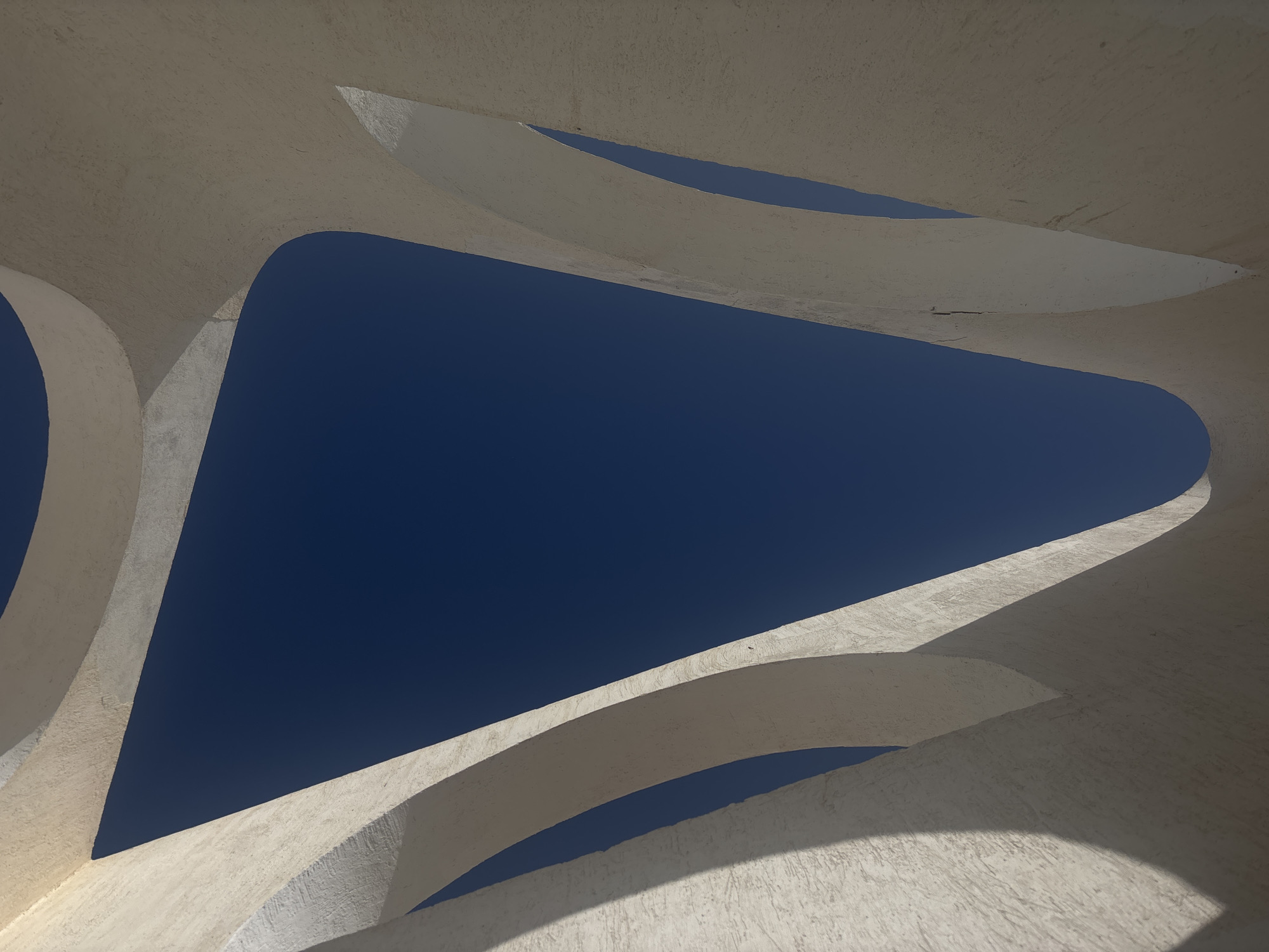 The image presents an architectural detail, likely an arch or structural support system, from an unknown building in an unknown city. The structures appear to be made of a light-colored, textured material, possibly concrete or plaster. The shapes are curved and interconnected, creating openings that reveal a deep blue sky. There are no people or identifiable objects beyond the architectural elements and the sky. The scene is well-lit, suggesting daytime. The overall impression is one of abstract design and minimalist composition.