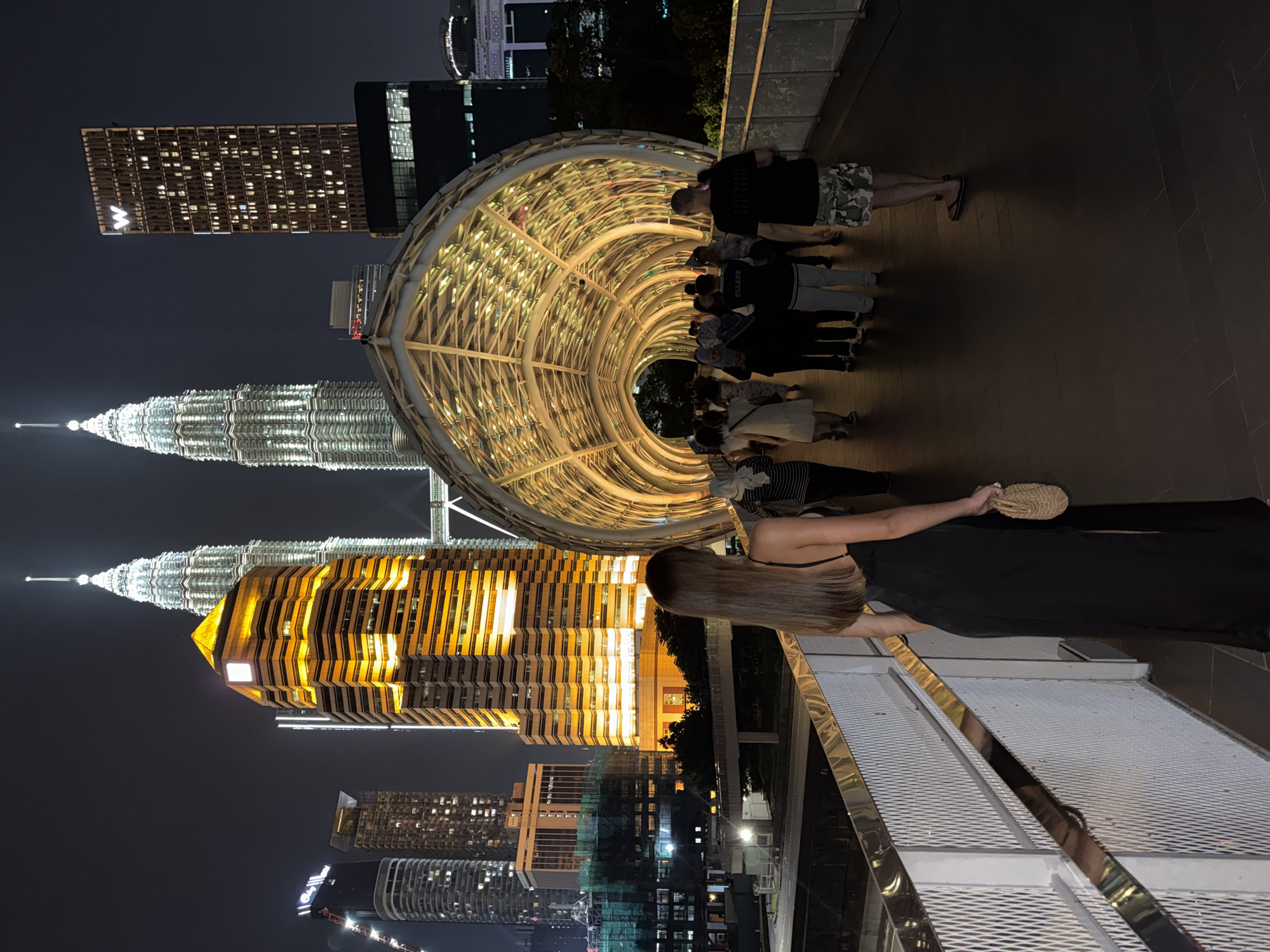 The image depicts a nighttime view in Kuala Lumpur, Malaysia. The Petronas Twin Towers are prominently visible, illuminated against the dark sky. A pedestrian bridge, featuring a distinctive arched structure with internal lighting, spans across the foreground. Several people are walking along the bridge. A woman in a black dress is standing on the bridge, facing the towers. Other buildings and structures are visible in the background, including a high-rise with illuminated windows.