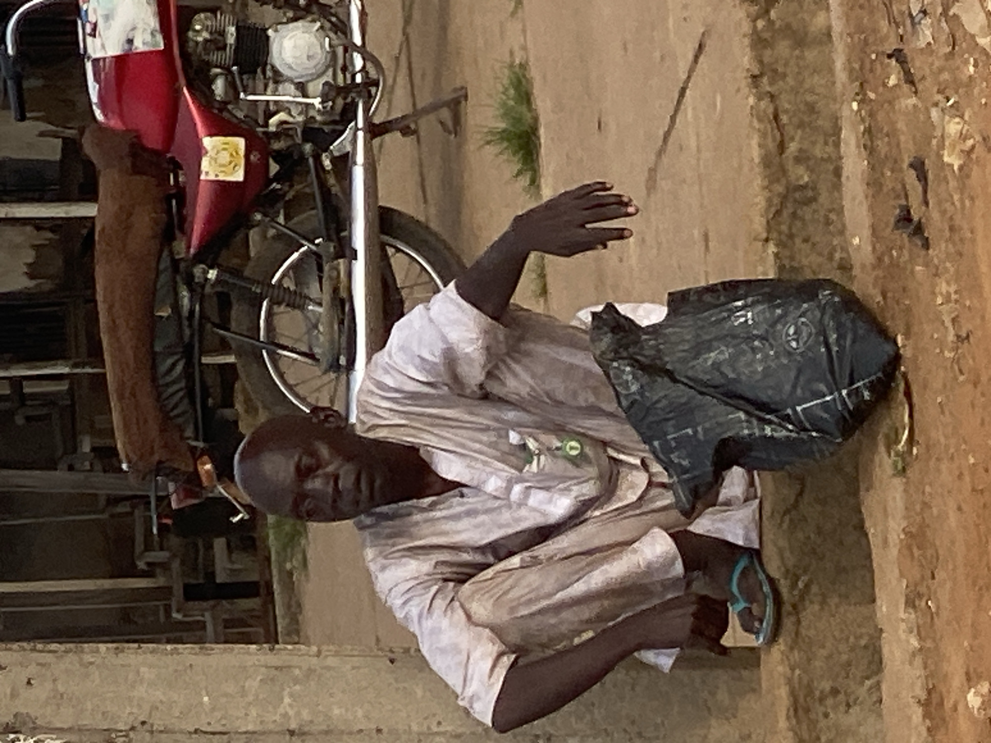 In Sabon Tasha, Nigeria, a man sits with his legs crossed on a raised concrete ledge. He is wearing a light-colored, loose-fitting traditional garment and blue flip-flops. To his right, a black plastic bag rests on the ground. Behind him, a red motorcycle with a brown seat is parked. The background shows a building with wooden structures. The ground is a mix of dirt and scattered debris.