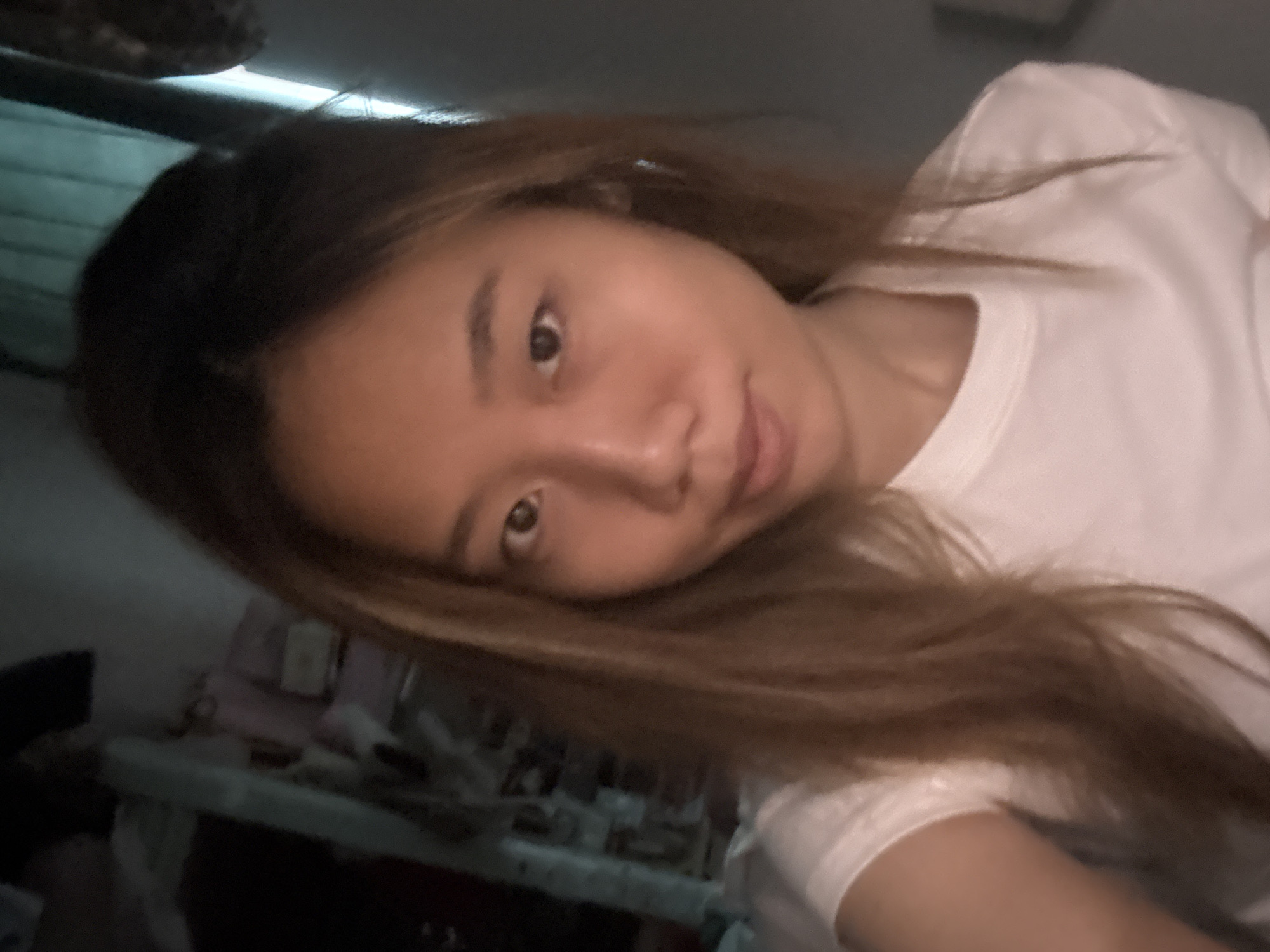 A woman with long, light brown hair is looking at the camera. She is wearing a white t-shirt. The background is dark and indistinct, but appears to be a cluttered room. The location might be deduced to be Nan'an District, China, based on implied information.