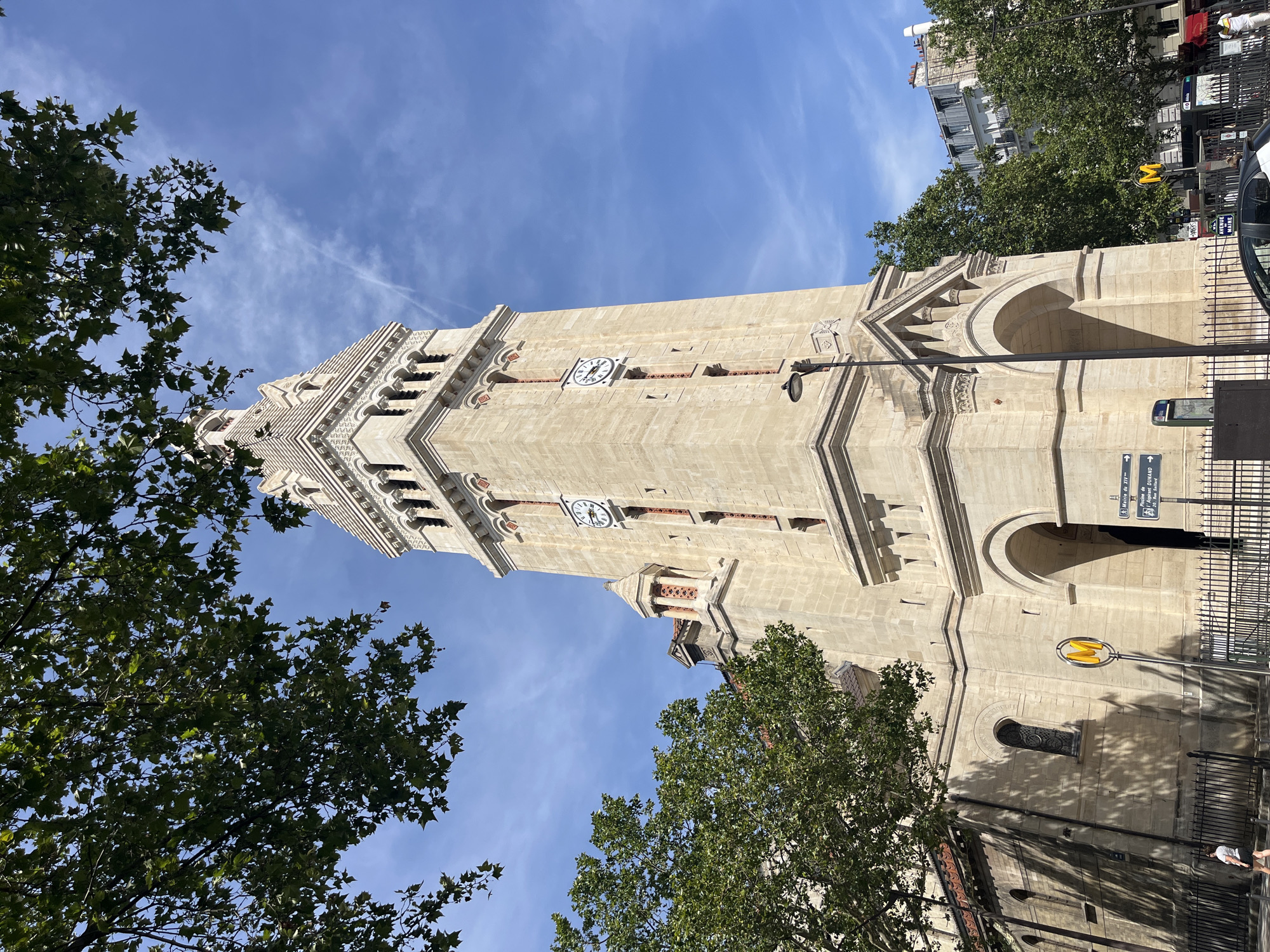 The image shows the exterior of the Église Sainte-Marguerite, a church located in the 11th arrondissement of Paris, France. The church features a tall, stone bell tower with a clock face. The tower has arched windows and decorative detailing near the top. Trees with green foliage are visible in the foreground, partially obscuring the sky and the upper parts of the church. The sky is blue with some scattered white clouds. In front of the church, there is a black metal fence. A yellow sign with a blue "M" symbol, indicating a Metro station, is visible on the left side of the image, along with street signs. A street lamp is positioned near the base of the tower. A few people are visible walking on the sidewalk in front of the fence. Buildings are visible in the background.
