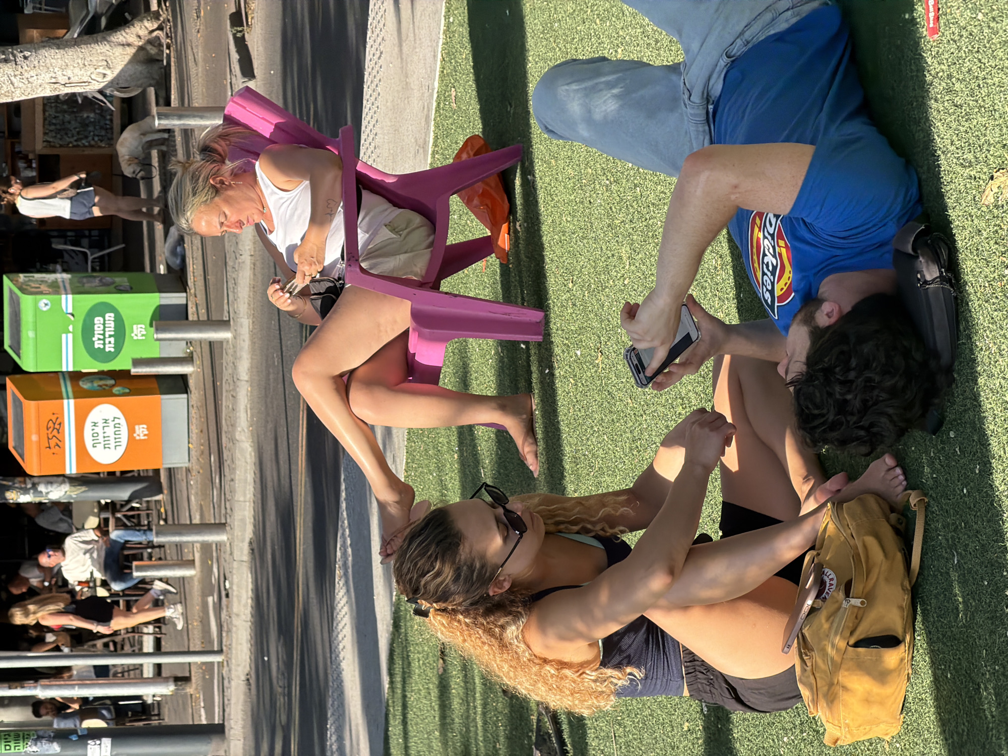 The image captures an outdoor urban scene in Tel-Aviv, Israel, featuring a public recreational area adjacent to a street and commercial establishments under bright daylight.
In the foreground, a patch of artificial green grass is occupied by three individuals. On the left, a young adult female with long, curly blonde hair and sunglasses is seated barefoot, wearing a dark blue tank top and black shorts. Her hands are positioned on her lap. Beside her, a mustard-yellow Kånken backpack is visible. To her right, a young adult male with dark curly hair lies on his back on the grass. He is wearing a blue t-shirt with a "Dickies" logo and light blue jeans, holding and operating a smartphone with his right hand. His head rests on a black fabric object. Further back on the artificial grass, a middle-aged female with blonde hair tied in a ponytail is seated barefoot in a bright pink plastic Adirondack-style chair. She is wearing a white tank top and light-colored shorts, and is actively engaged with a smartphone held in both hands. An orange plastic object is partially visible underneath her chair.
Beyond the grass area, an asphalt street with white markings runs horizontally. Across the street on a concrete sidewalk, various urban elements are present. These include an orange recycling bin with Hebrew text "איסוף אריזות למחזור" (Collection of packaging for recycling) and a green mixed waste bin labeled "פסולת מעורבת" (Mixed waste). Multiple silver bollards are positioned along the sidewalk. Further into the background, several individuals are seated at outdoor tables of a cafe or restaurant, under dark awnings. One man in a white t-shirt is clearly visible among them. A woman in a white top and dark shorts is seen walking with a light-colored dog on a leash near a tree trunk on the right side of the frame. The scene is well-lit by direct sunlight, creating distinct shadows.
