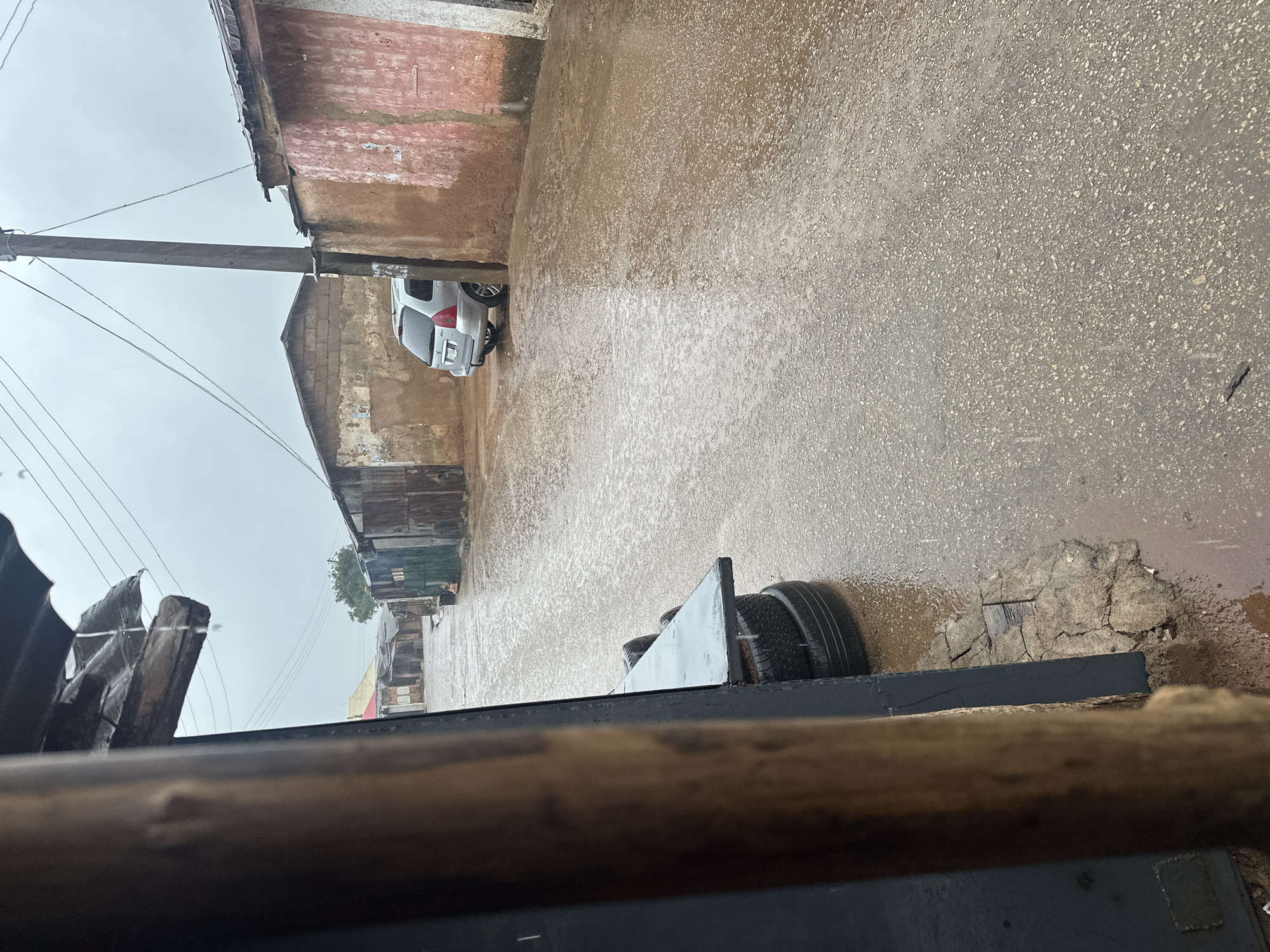 The image depicts a street scene in what appears to be an urban or semi-urban environment, characterized by rain. The foreground is dominated by the edge of a dark structure on the left, with a rough, wooden beam protruding from above. The street itself is wet and appears to be covered in shallow puddles, indicating recent or ongoing rainfall. Several corrugated metal structures and buildings with earthy-toned walls line the sides of the street. A silver hatchback car is parked on the right side of the street, partially obscured by a wall. In the distance, more buildings and trees are visible under a cloudy sky. A utility pole with multiple wires is also present. On the left side of the street, a stack of tires is visible, supporting a flat surface. The overall atmosphere is somber due to the overcast weather and the rain.