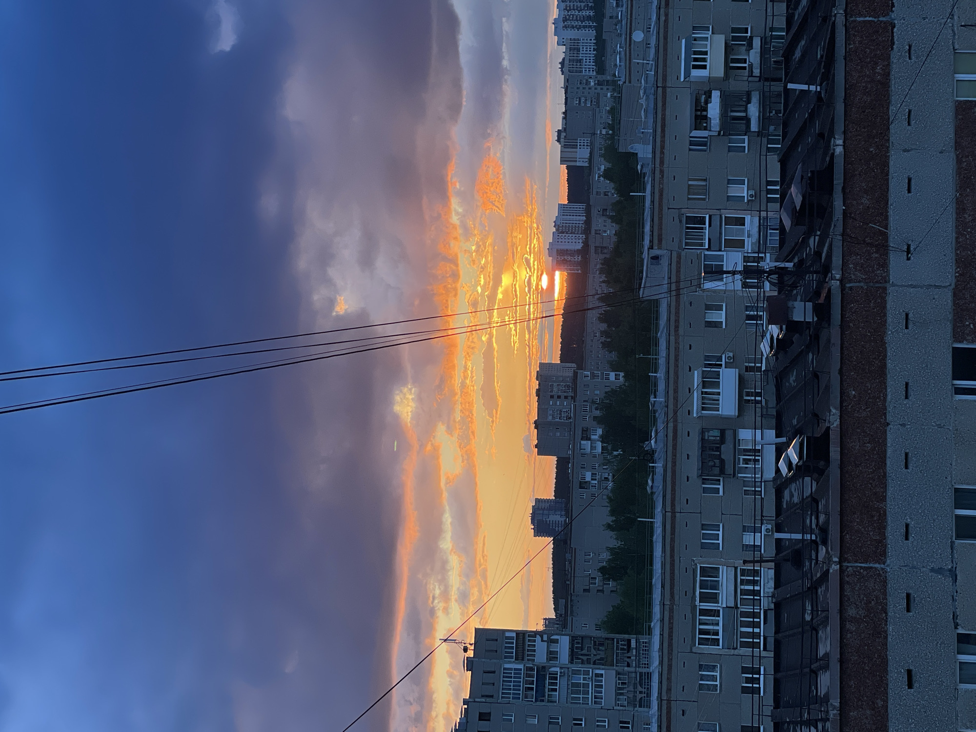The photograph depicts a sunset over a cityscape, likely taken from an elevated position. The lower half of the frame is dominated by buildings, typical of urban residential architecture in the city of Izhevsk, Russia. These are predominantly grey apartment blocks, with visible windows and balconies. A darker, vertical band with brick detailing runs along the right edge of one building.
The upper half of the image features a dramatic sky with a gradient from a dark blue on the left to orange and yellow hues near the center where the sun is setting. Clouds are streaked across the sky, reflecting the colors of the sunset.
Power lines are visible in the lower left, cutting across the sky. The overall scene is urban, with the focus on the contrast between the built environment and the natural beauty of the sunset. The light is dim and appears to be capturing the twilight. There are no people visible in the image.
