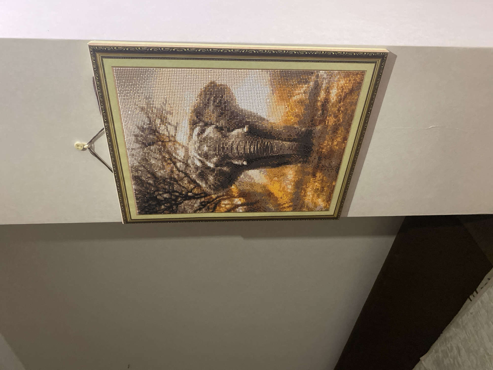 The image shows a diamond painting of an elephant in a savanna landscape, framed and hanging on a pale beige wall. The painting is positioned in the upper right quadrant of the image. The elephant is depicted centrally, facing forward, with a large tree behind it and a sunlit, grassy terrain. The frame is ornate with a gold and dark green pattern. Below and to the left of the painting, a dark brown headboard of a bed is visible, with a white, textured bedspread in the foreground. The overall lighting suggests an indoor setting. There is no discernible information in the image that would allow for identification of its location in Tolyatti, Russia.