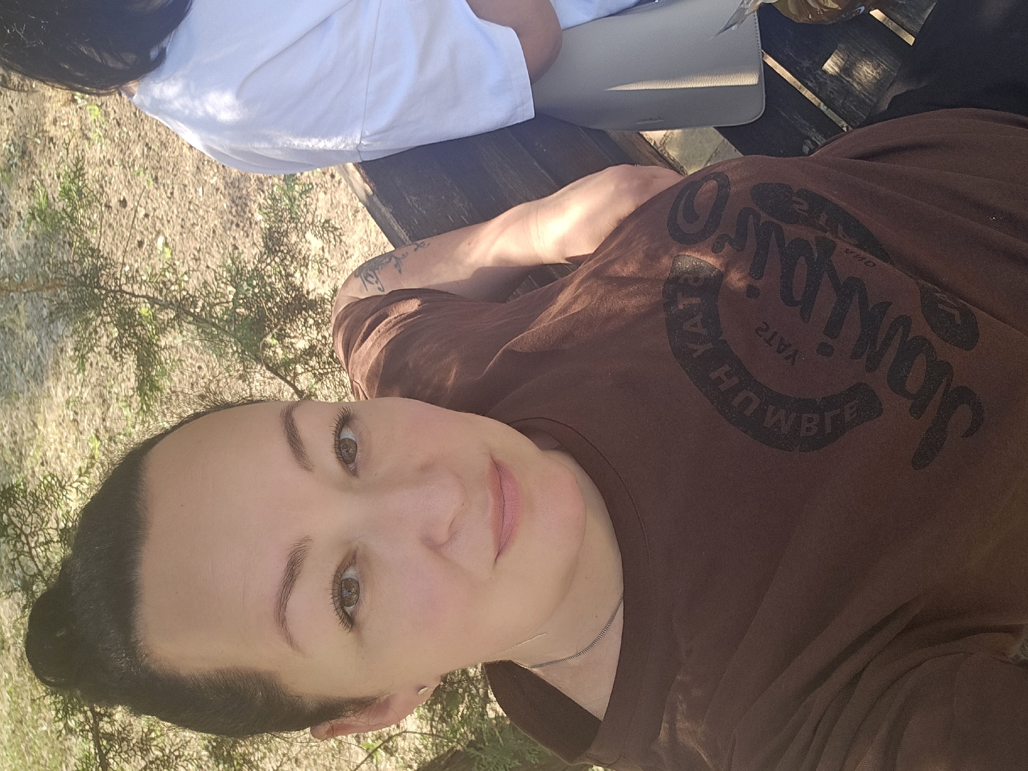 The image shows a close-up of a person's face, oriented sideways. The person has dark hair pulled back, brown eyes, and is wearing makeup, including pink lipstick. They are wearing a brown t-shirt with a dark graphic print. The background shows a person in a white shirt and a wooden surface, possibly a table. The setting appears to be outdoors, with dry ground and some greenery. There is a tattoo visible on the arm of the person in the brown shirt, and text on the t-shirt appears to be "Jamming" and "Katya". The lighting suggests it is daytime, with sunlight casting shadows. The overall mood is casual and relaxed. This image was taken in Skadovsk, Ukraine.