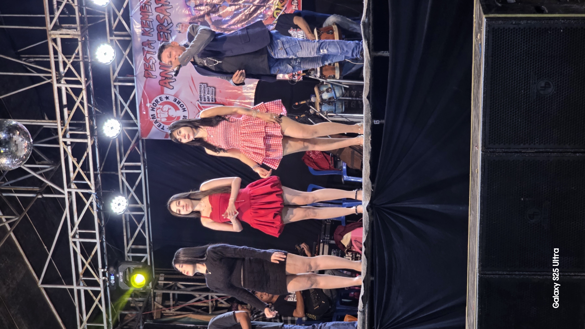 A medium shot captures a group of people on a stage in Juwana, Indonesia. Three women stand in the foreground, all wearing short dresses. The woman on the far left wears a black long-sleeved dress, while the middle woman is in a red sleeveless dress, and the woman on the right wears a red and white checkered dress. A man stands to the right of the women, holding a microphone and wearing a blue blazer over a dark shirt and ripped jeans. Behind them, a banner displays text and graphics. The stage is lit by bright spotlights and a disco ball hangs above. In the background, musical instruments, including drums, and speakers are visible. The setting appears to be an outdoor event at night.