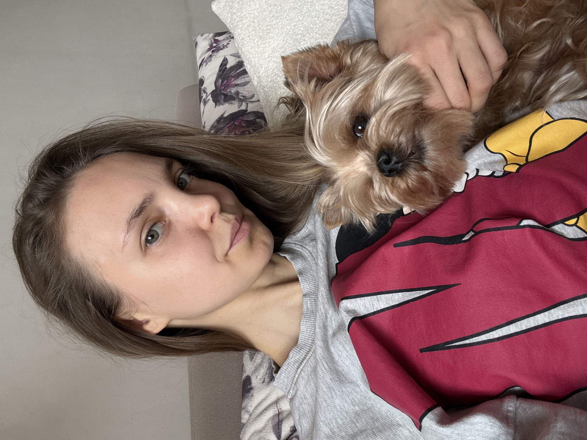 A woman with long, brown hair and fair skin lies on a bed, with a small, brown dog nestled close to her chest. The dog appears to be a Yorkshire Terrier. The woman is looking at the camera with a neutral expression. She is wearing a gray, long-sleeved shirt with a large, red and yellow cartoon design on the front. Her right hand rests on the dog. The bed has a patterned pillow behind the woman's head and a light-colored headboard. The wall behind the bed is a neutral color. The location of Tbilisi, Georgia is not discernable from the visual information provided.