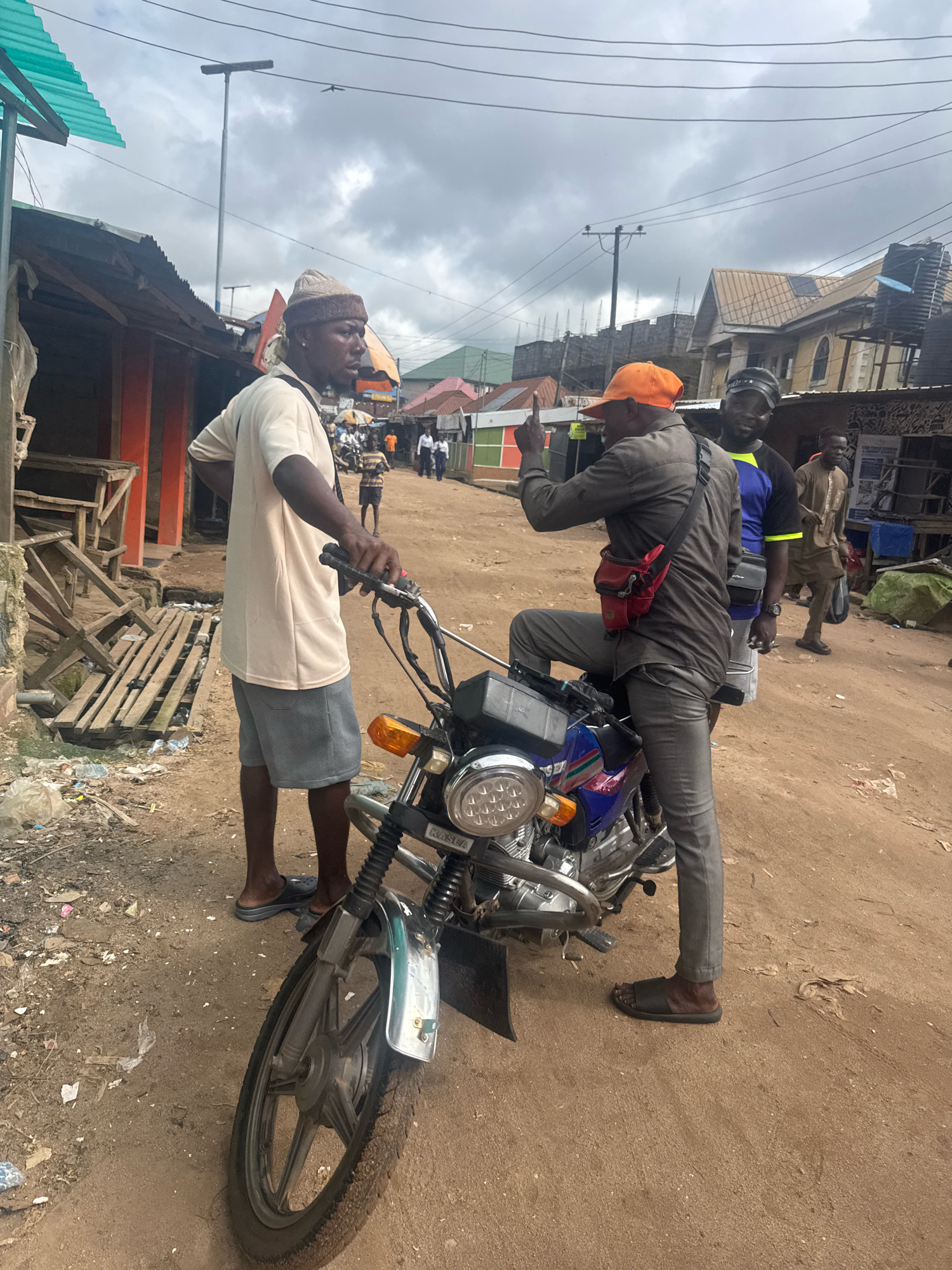 In Ihiagwa, Nigeria, an outdoor scene unfolds under a cloudy sky. Two men are engaged in conversation near a parked motorcycle. One man, wearing a cream-colored t-shirt, gray shorts, and a knit cap, stands beside the motorcycle. The other man, dressed in a gray long-sleeved shirt, gray pants, and an orange cap, is seated on the motorcycle, gesturing with his right hand. A third man, wearing a blue and yellow t-shirt, stands behind the man on the motorcycle. In the background, several other individuals are visible, along with buildings and market stalls. The ground is covered in dirt and debris, typical of an unpaved urban area.