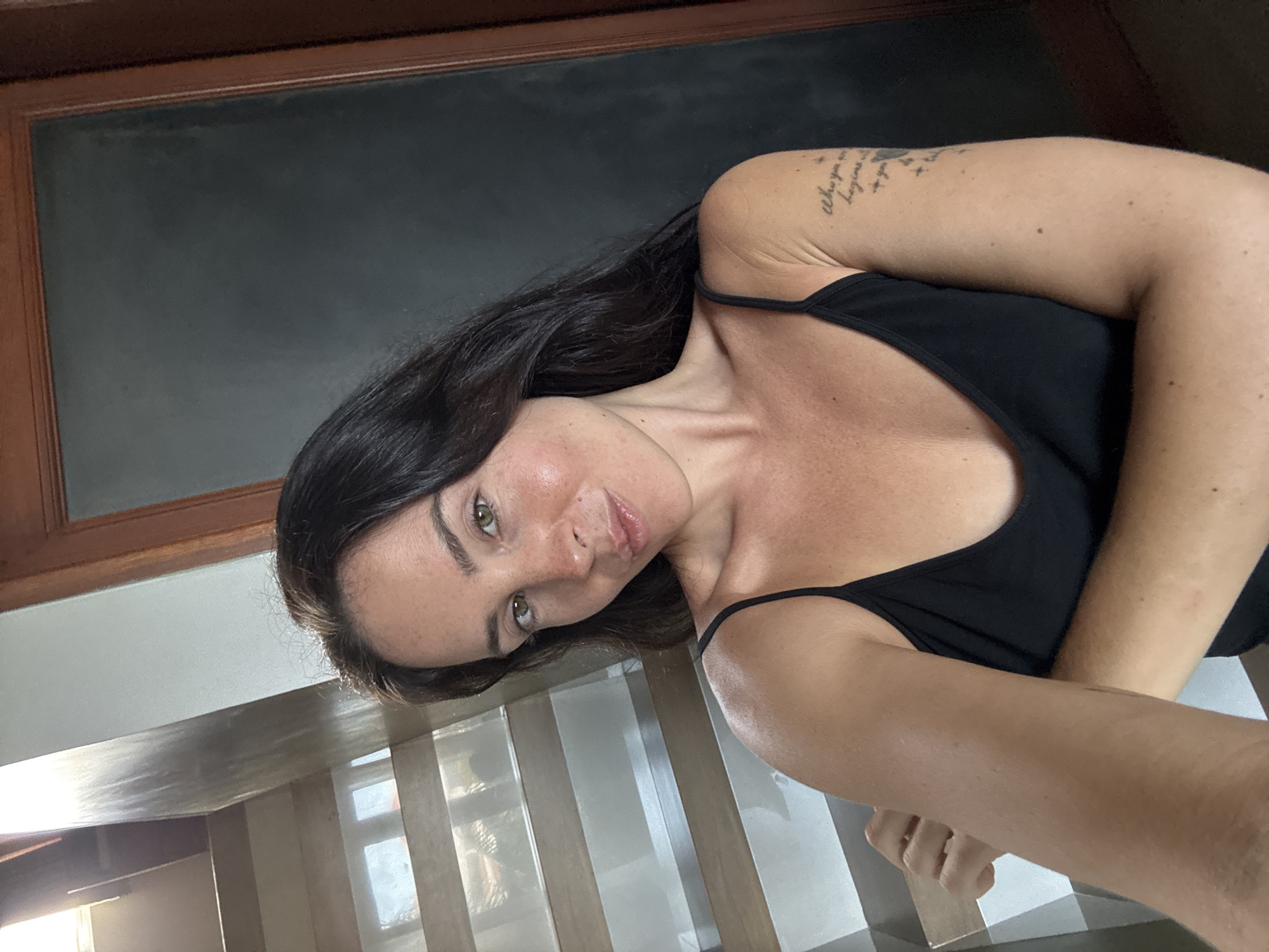 A young woman with dark hair and fair skin, wearing a black tank top, poses for a selfie in what appears to be an indoor setting with a staircase to her left and a dark-colored door to her right. Her right arm is extended towards the camera. A tattoo is visible on her upper left arm. The environment is well-lit, suggesting it could be daytime. The image does not contain any explicit visual cues to confirm its location within North Kuta, Indonesia.