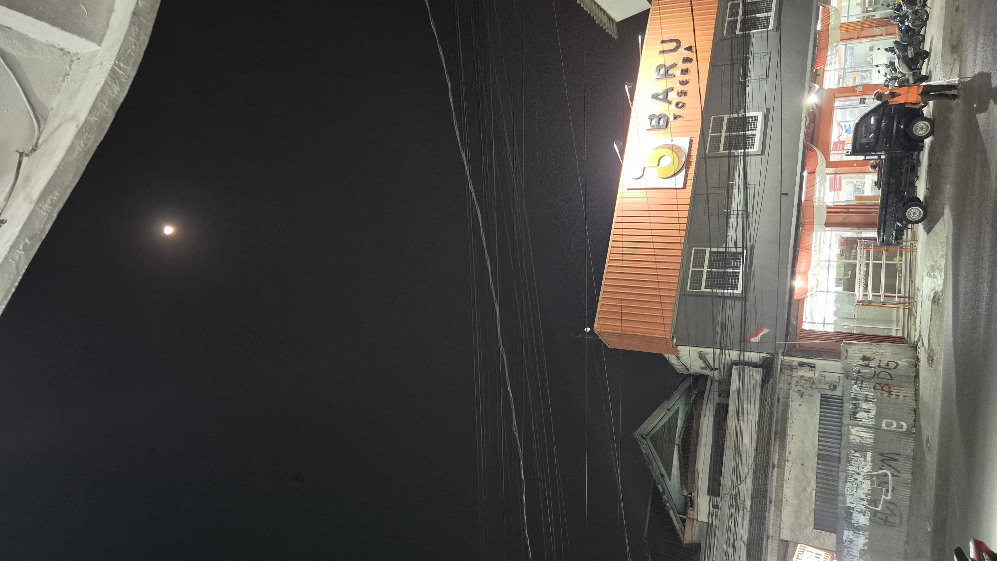 This is a nighttime photograph taken from a street level perspective, looking upwards towards a building facade. The sky above is dark and appears to have a reddish-orange celestial body, possibly the moon, in the upper quadrant. Several electrical wires crisscross the sky.

Dominating the middle ground is a commercial building with an orange corrugated metal facade on its upper level. Below this, the building has a gray lower section with several windows. A sign above the entrance reads "BARU TOSERBA". A small Indonesian flag is attached to a pole on the left side of the building. The entrance to the building is open, revealing shelves and displays inside.

In front of the building, a black pickup truck is parked. A person wearing an orange safety vest stands to the right of the truck, holding a long pole. Several motorcycles are parked to the right of the truck.

To the left of the commercial building, there are other structures. One has a green-tiled roof and a lighter colored facade with corrugated metal siding. Graffiti is visible on the metal siding.

The street in the foreground is asphalt and appears wet, reflecting some of the ambient light. The overall scene suggests a commercial area in Karawang, Indonesia, captured after dark.
