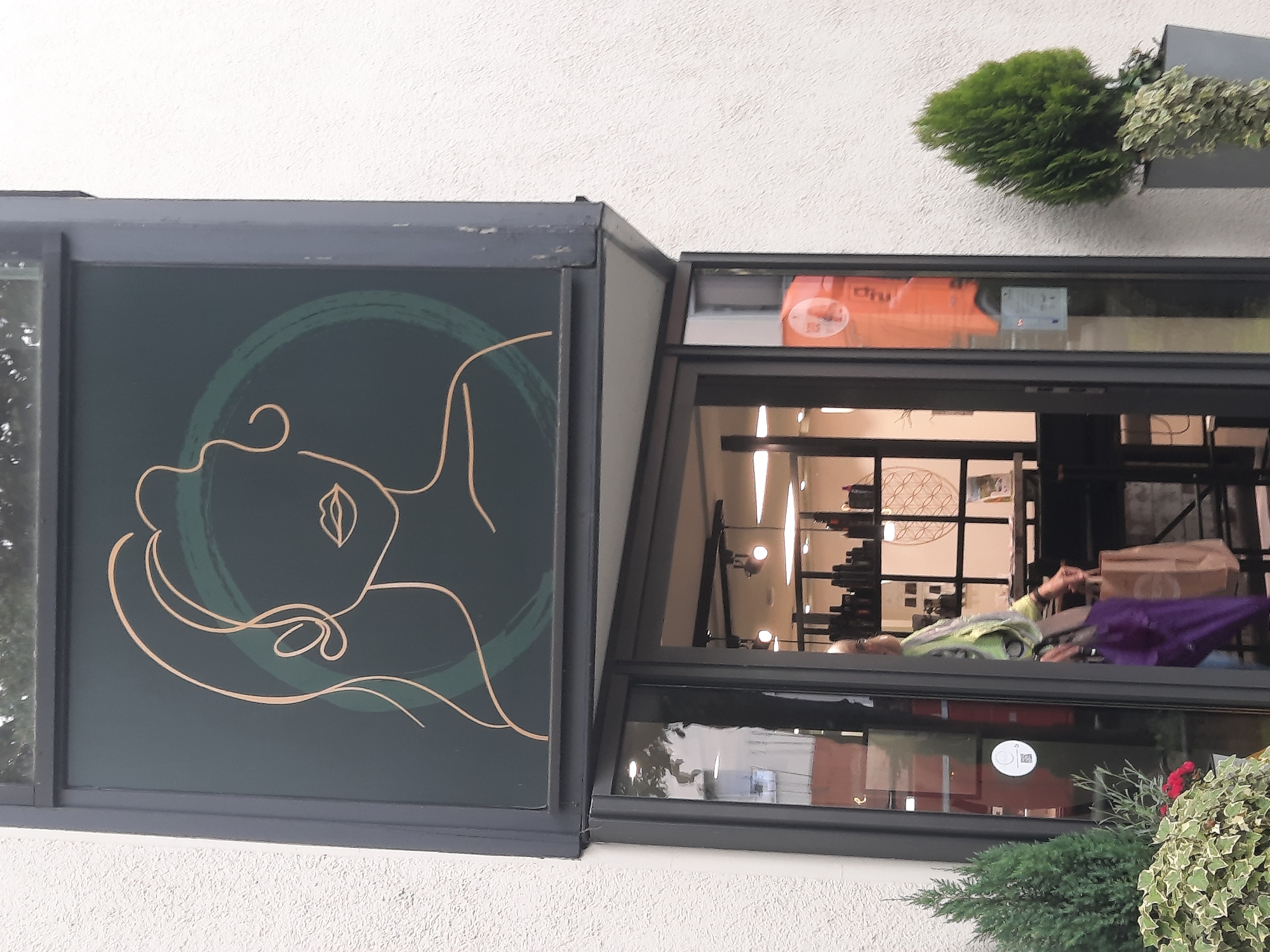 A storefront is pictured in Rosenheim, Germany, featuring a dark green sign with a minimalist line drawing of a woman's profile in gold and green. Below the sign, a glass door leads into the establishment, which appears to be a salon or beauty shop, with shelves of products visible inside. A woman is entering the shop, carrying a purple umbrella and a brown paper bag. She is wearing a light green, patterned scarf and brown pants. In front of the store, there are two potted evergreen plants, one on the left and one on the right. The building's exterior wall is white and textured.
