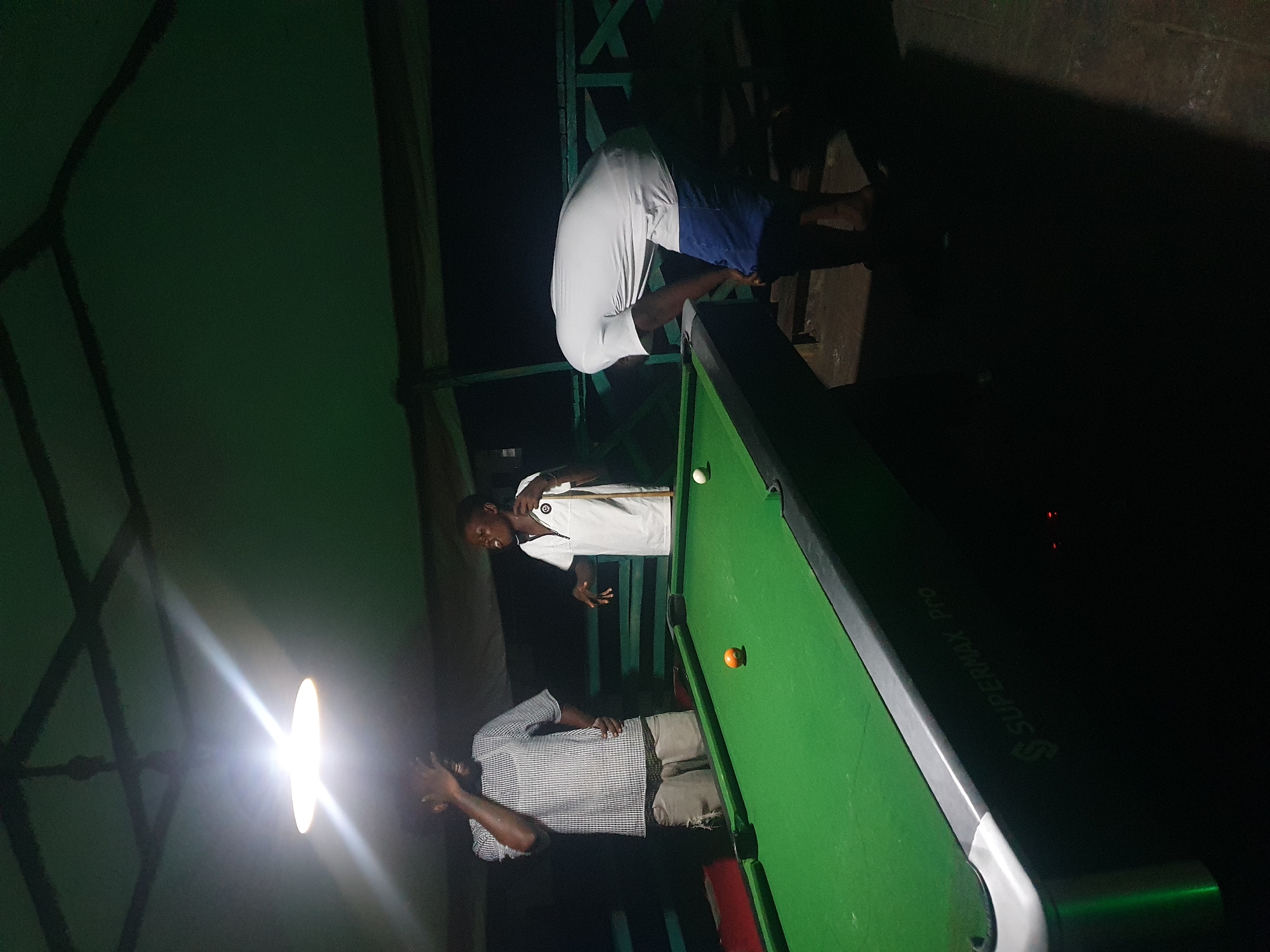 A group of young men are playing pool at night under a covered outdoor area, possibly a bar or recreational spot in Lokoja, Nigeria. The scene is dimly lit by a single overhead light fixture, casting harsh shadows. In the foreground, a green pool table with two balls, one orange and one white, is visible. On the left, a man in a checkered shirt and ripped shorts stands by the table, one hand on his head. To his right, another man in a white t-shirt with a zipper and a gold emblem is holding a pool cue, looking animatedly towards the other side of the table. On the far right, a third man, wearing a white t-shirt and blue shorts, is bent over the pool table, seemingly preparing to take a shot. The background shows a dark, possibly wooden, structure with railing, suggesting an open-air setting. The overall atmosphere is casual and social, with the men engaged in a game of pool.