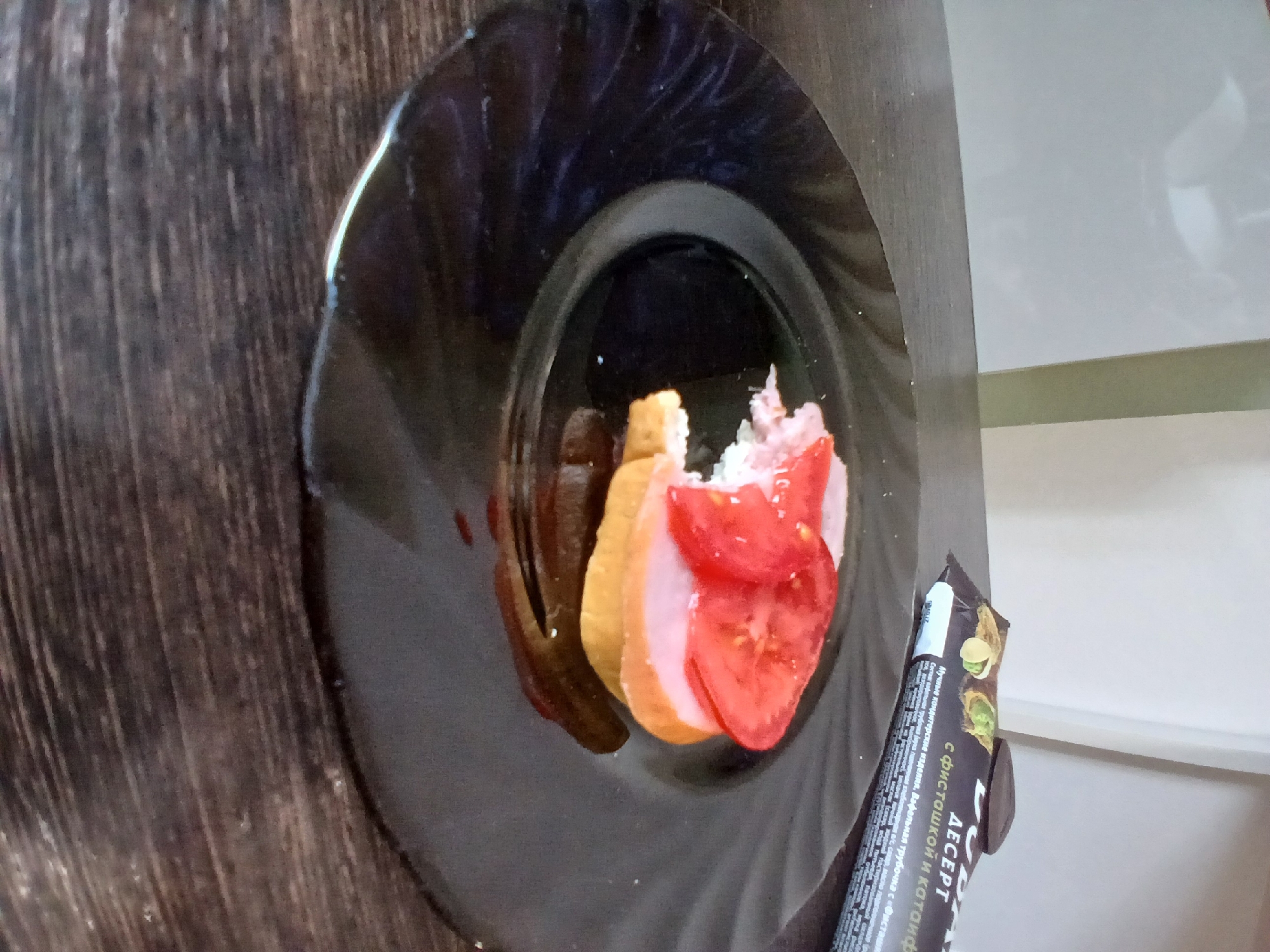 A sandwich is on a black plate. The sandwich has bread, ham, and tomato. There is also a dark brown wrapper with text that says "DESSERT" in Russian. The scene is indoors, with a wooden table and a light-colored wall in the background.
