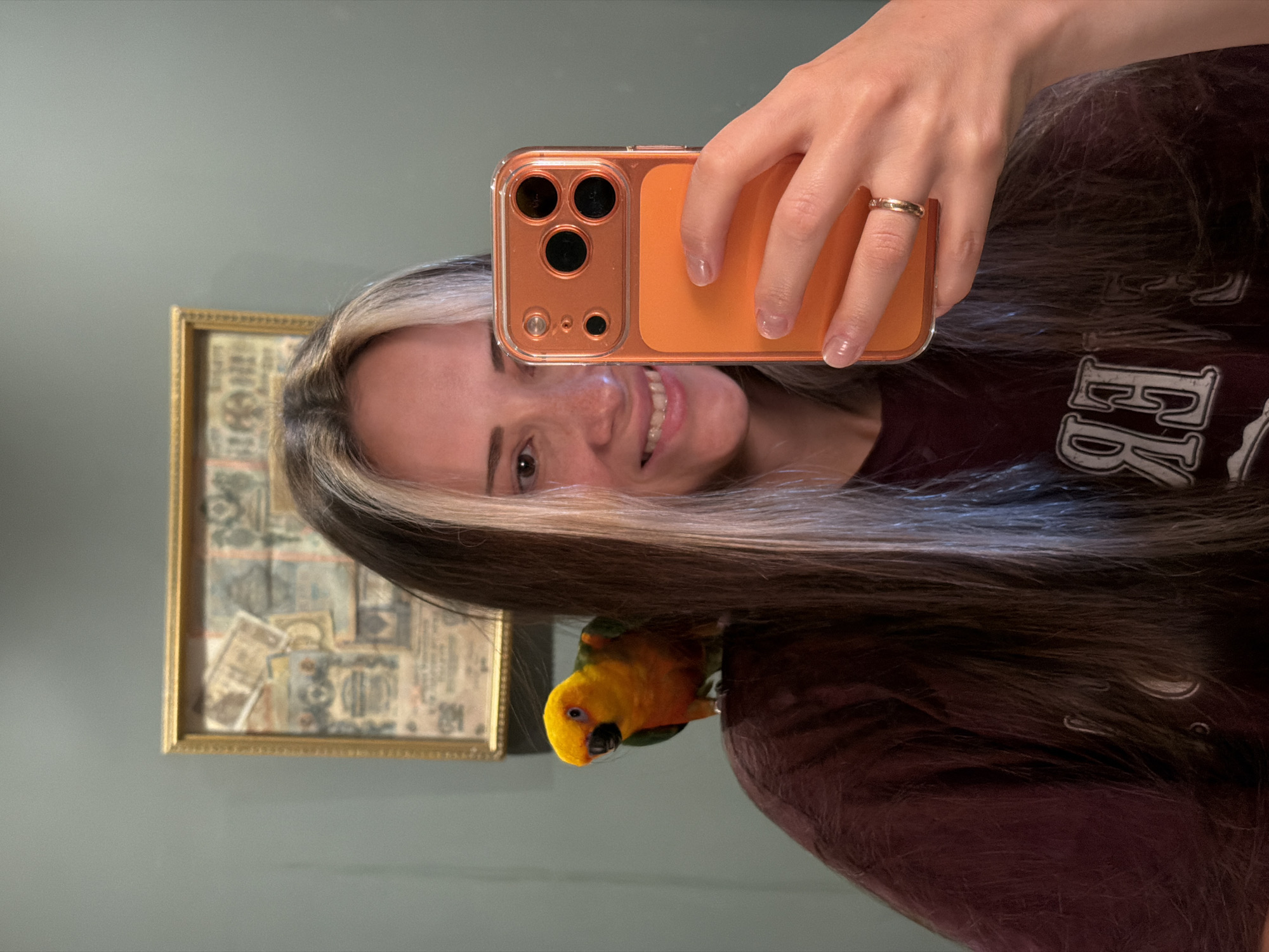 A person is taking a selfie in a mirror. They are holding an orange iPhone in their right hand. The person has brown hair with blonde streaks. A yellow and green bird is perched on their left shoulder. Behind the person, a framed picture hangs on a gray wall. The person is wearing a dark maroon shirt. There is a ring on the person's finger. The location is not identifiable as Khimki, Russia from the information in the image.