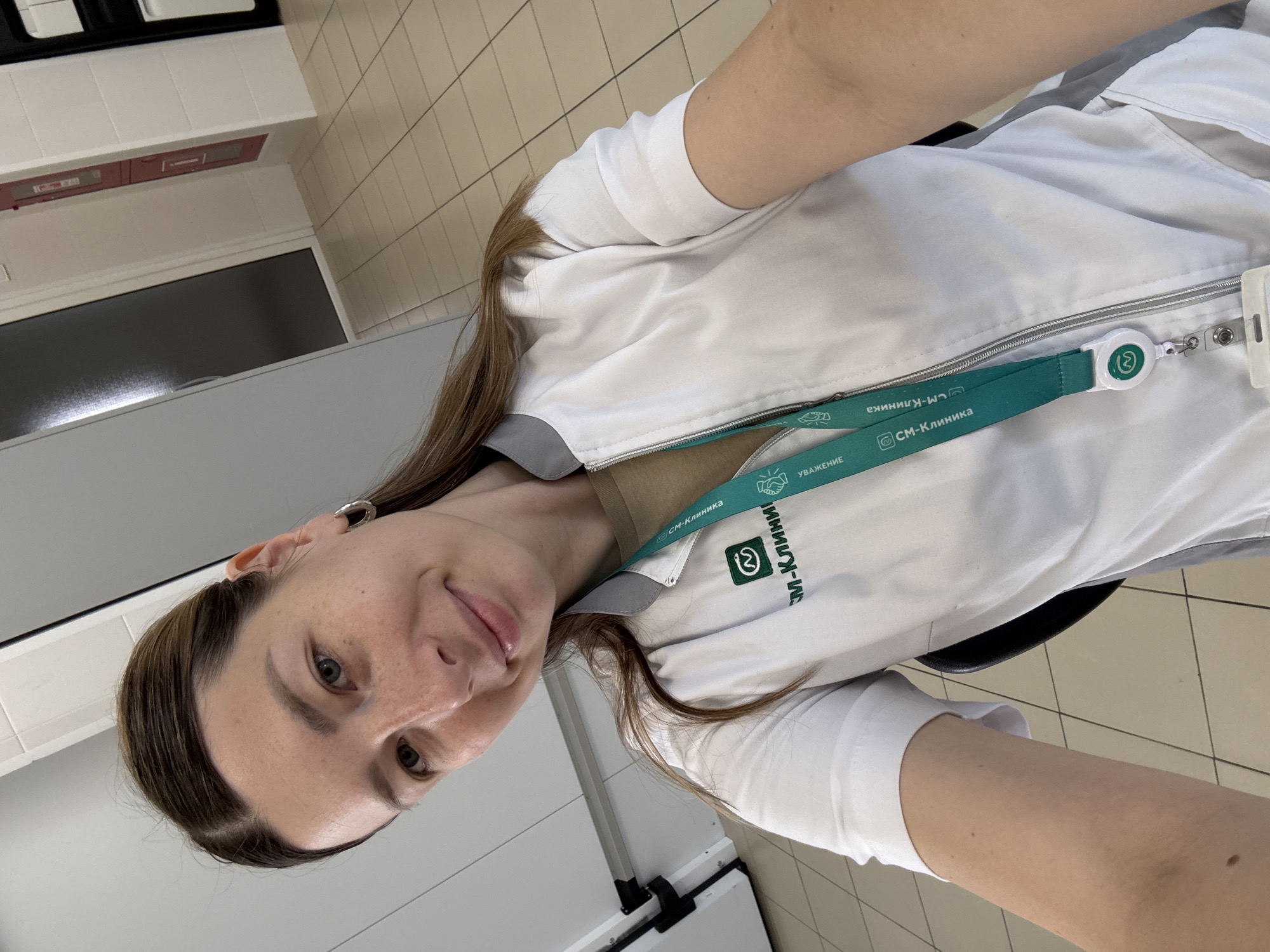 A woman is taking a selfie in a medical facility. She is wearing a white lab coat with a green lanyard. The lab coat has a logo that reads "СМ-Клиника". The background appears to be a hallway with tiled floors and white walls, suggesting it is an indoor medical setting. The woman is looking at the camera and smiling. The scene is brightly lit. The general appearance of the hallway, including the flooring and wall finishes, is consistent with modern clinic interiors found in Moscow.