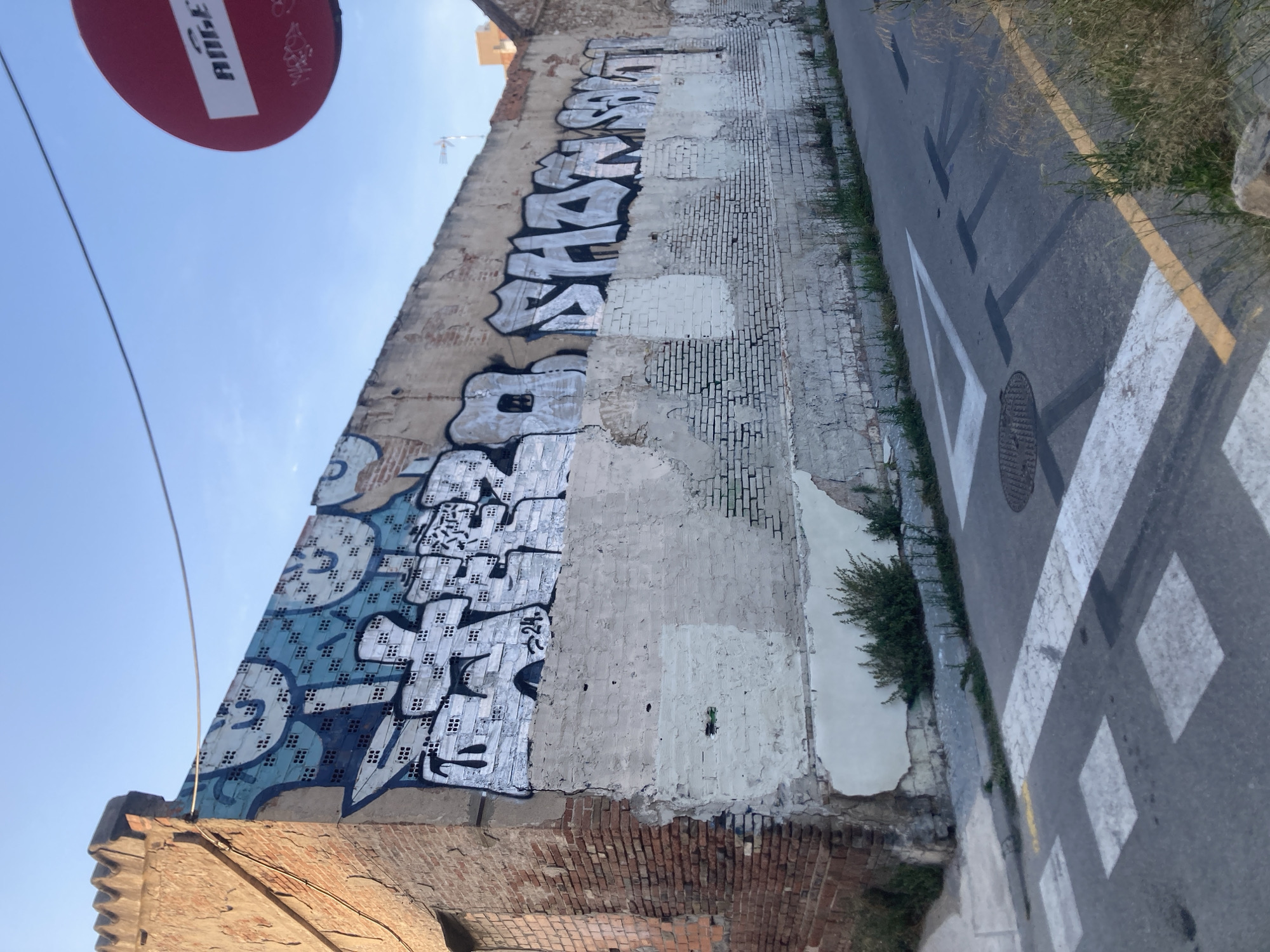 The image shows an urban street scene in Barcelona, Spain. A dilapidated brick building on the left is covered in graffiti, with visible paint peeling and crumbling plaster. The graffiti features large, stylized lettering in black, white, and blue, some with intricate patterns. The wall extends into the background, where more graffiti is present. A red circular traffic sign with white lettering that appears to say "ROGER" is visible in the upper right corner, also tagged with graffiti. The street in the foreground has a pedestrian crossing marked with white stripes and a yellow line. The road surface shows painted road markings, including a triangle and what appear to be tire marks casting shadows. There are no people visible in the image. The environment suggests an area with some neglect, characterized by the decaying building and street art. The sky is clear and blue, indicating daytime.