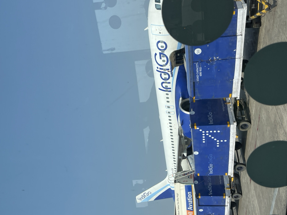 The image, rotated 90 degrees clockwise, captures an IndiGo airline aircraft on the tarmac of an airport, likely in Guwahati, India, during bright daylight. The main subject is a white airplane with blue livery, featuring the prominent "IndiGo" logo along its fuselage. The aircraft's registration number, "VT-ITF," is visible near the tail.
Adjacent to the aircraft, several blue cargo containers or baggage carts are positioned, engaged in ground operations, suggesting either loading or unloading. These containers are also branded with "IndiGo CarGo" text and display the airline's stylized 'i' logo (which resembles an arrow). One container clearly states "LOAD CAPACITY 600 KGS" and the number "58." A set of mobile stairs or a ramp is connected to a door on the aircraft, indicating passenger boarding or deplaning activity.
Further text visible on the aircraft's tail section includes "IndiGo" and a partial slogan that reads "Work Takes You Sky High." Another partially visible text reads "il Aviation," possibly part of "GoIndiGo Aviation." The scene is viewed through a window, as evidenced by the reflections of dark circular objects (likely camera artifacts or interior elements) on the glass and the slightly tinted blue hue of the upper portion of the image, representing the sky. The weather appears clear and sunny. No people are distinctly visible in the frame.