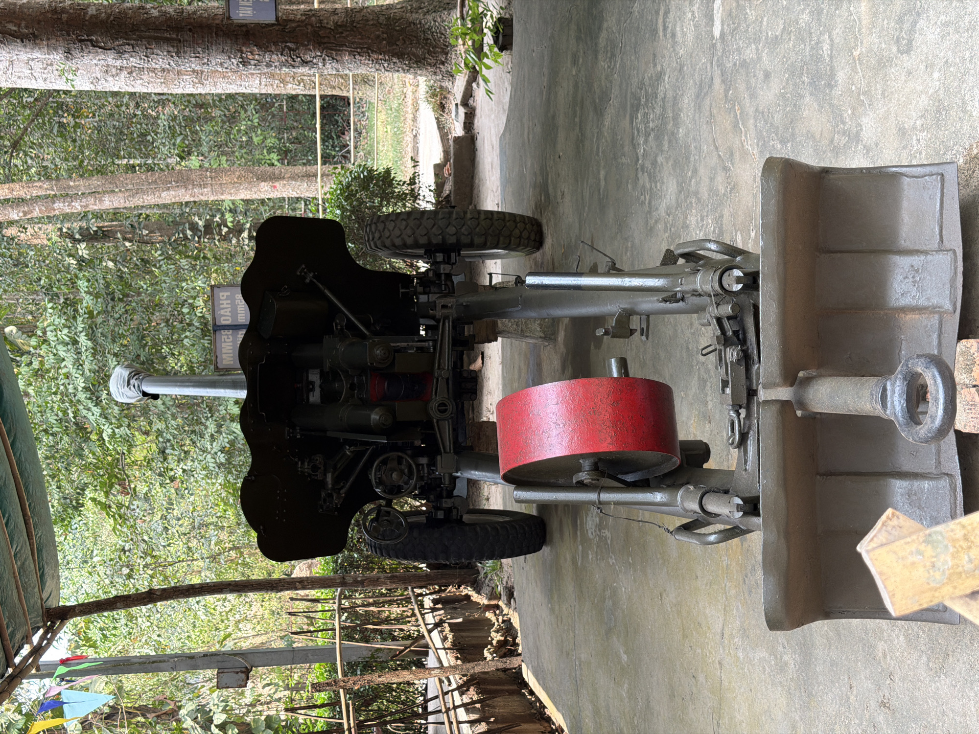 The image displays a large, grey-green artillery piece or heavy weapon positioned outdoors on a concrete surface. The weapon features a prominent long barrel, two robust rubber-tired wheels on either side of its carriage, and an assortment of mechanical parts including gears, levers, and a distinct red cylindrical component, possibly a counterweight or protective cover, centrally located. To the right of the main weapon, a heavy, flat, shovel-shaped base plate or ground anchor is visible.
The setting appears to be a historical display area, with a backdrop of lush, dense tropical foliage and tall trees, suggesting a natural environment surrounding the exhibit. The ground beneath the weapon is concrete. The scene is brightly lit by natural daylight, indicating a clear, sunny day, likely midday, with strong shadows visible on the ground. There is no activity taking place; the weapon is static, presented for viewing.
In the upper left portion of the image, partially obscured by trees, a dark sign is visible. The text on the sign is reversed, but when mentally inverted, parts of it read "ĐẠN HOẠ" (likely Vietnamese for "artillery shell" or similar) and the clear numerical designations "MM28" and "MM32", which probably refer to the weapon's caliber or ammunition types. The location context of Ho Chi Minh City suggests this could be part of a war museum or memorial.