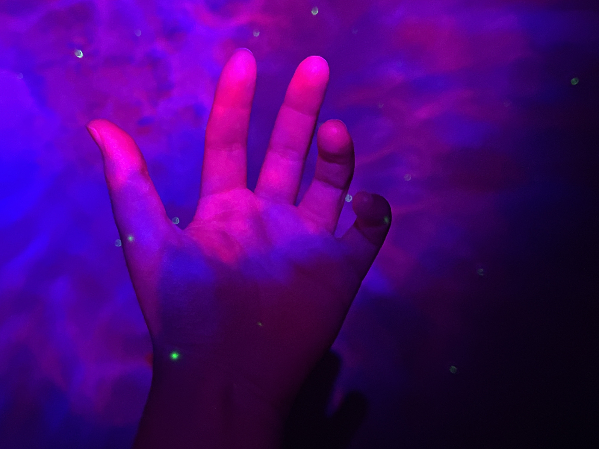 The image shows a close-up of a person's hand, illuminated by purple and pink light. The hand is open, with fingers slightly spread. Several small, bright green specks are visible on the hand and in the surrounding atmosphere. The background is a hazy, abstract blend of purple and blue, suggesting a smoky or atmospheric effect. The lighting creates a dramatic and somewhat ethereal mood. There is no discernible location in Saint Petersburg, Russia, or any other specific urban environment depicted. The scene is abstract and focused on the visual elements of light, color, and texture on the hand.