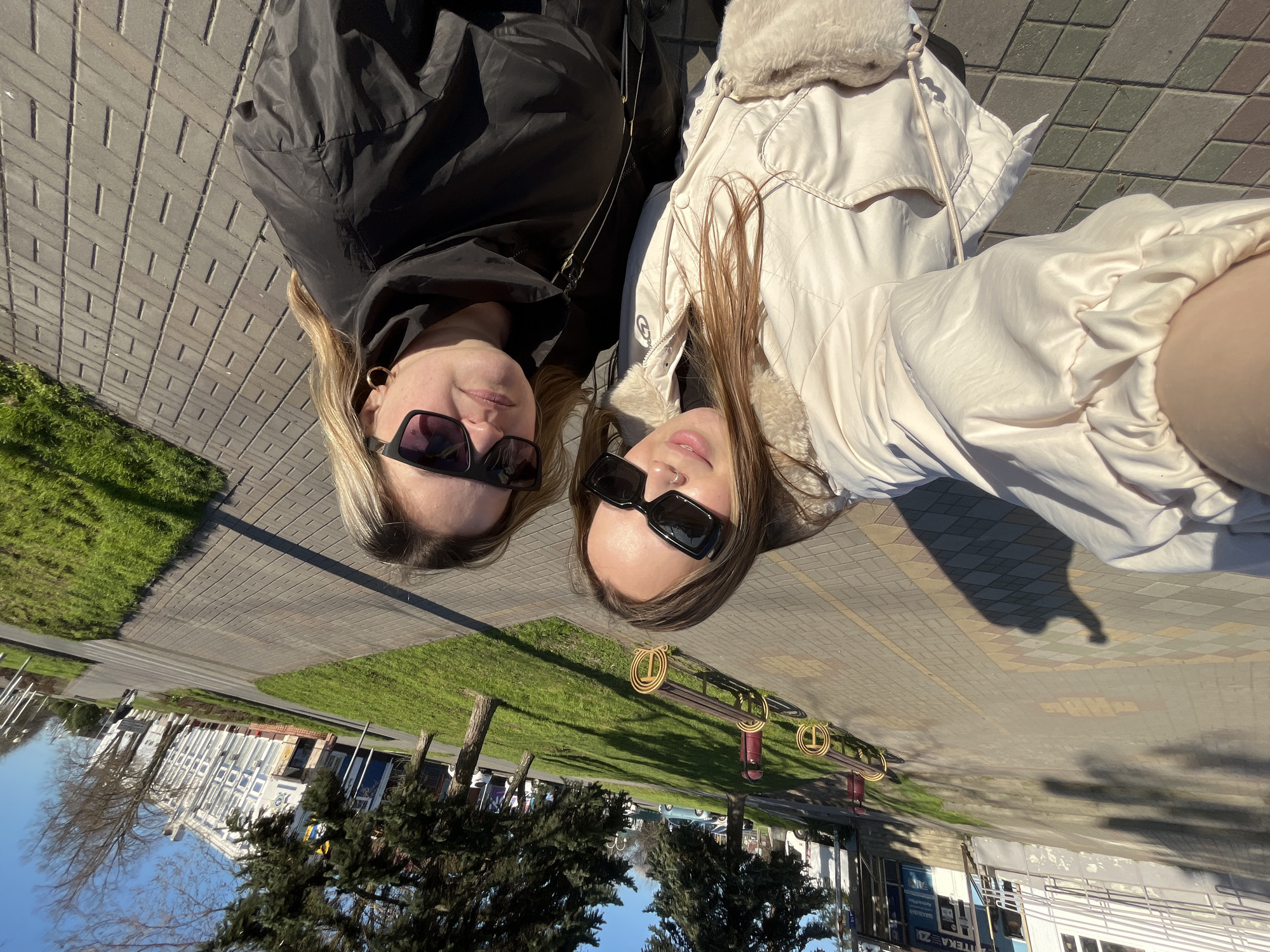 Two women are positioned lying down on a brick surface, likely a pedestrian walkway or square. They are wearing sunglasses. One is wearing a black jacket. The other is wearing a light-colored jacket with a fur-lined collar. The background shows a mix of green grass, trees, and buildings. A street sign is visible. Based on the text on the sign in the background, the location is Novopokrovskaya, Russia. A bench with ornate metal details is in the background as well.
