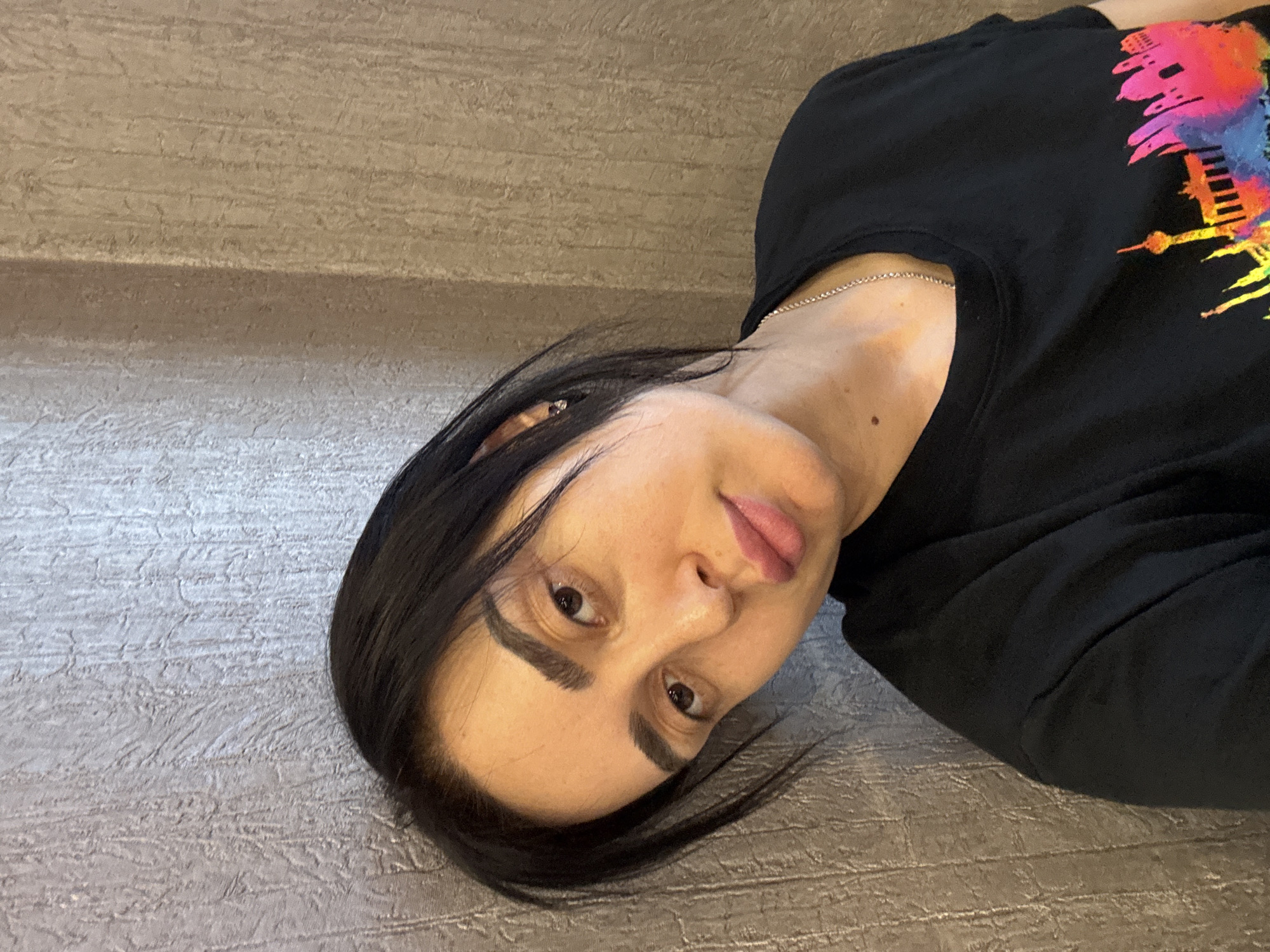 A woman lies on a textured gray surface. She has dark hair and is wearing a black t-shirt with a colorful skyline design, including a recognizable structure that resembles the Tbilisi TV Broadcasting Tower. She is also wearing a silver necklace. Her skin is fair and she has dark eyebrows. The background appears to be a wall with a textured finish. The image strongly suggests that the skyline design on the woman's shirt relates to Tbilisi, Georgia, as the TV tower is a prominent landmark there.