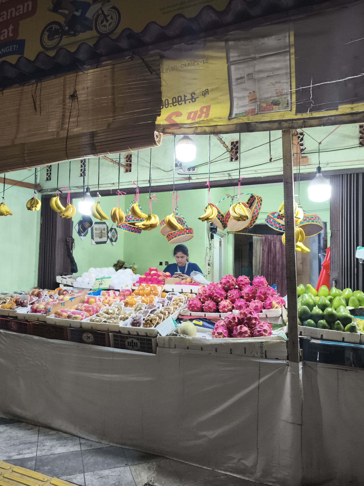 The image captures a nighttime scene at a fruit stall in Depok, Indonesia. A vendor, a woman wearing a blue top, is positioned behind a counter laden with a vibrant assortment of fruits. She is looking down at her phone, seemingly engaged with it while working.
The stall is well-stocked, displaying a variety of produce including bunches of bananas hanging from the ceiling, stacks of pink dragon fruits, numerous green avocados, and an array of other fruits packaged in clear plastic containers. The fruits are arranged on tiered displays, with white styrofoam boxes and plastic crates used for presentation and organization. Decorative, woven hats with colorful patterns are also hanging from the ceiling, adding a festive touch to the stall.
The stall is illuminated by several bright, bare light bulbs hanging from wires, casting a direct and somewhat harsh light on the scene. The background shows a light green wall. Above the stall, a large banner with images of a motorcycle and a refrigerator is visible, along with some text that is partially obscured and difficult to read. The overall environment appears to be an outdoor market or roadside stall, with a paved ground visible in the foreground. The image is taken from a slightly elevated perspective, looking towards the stall.