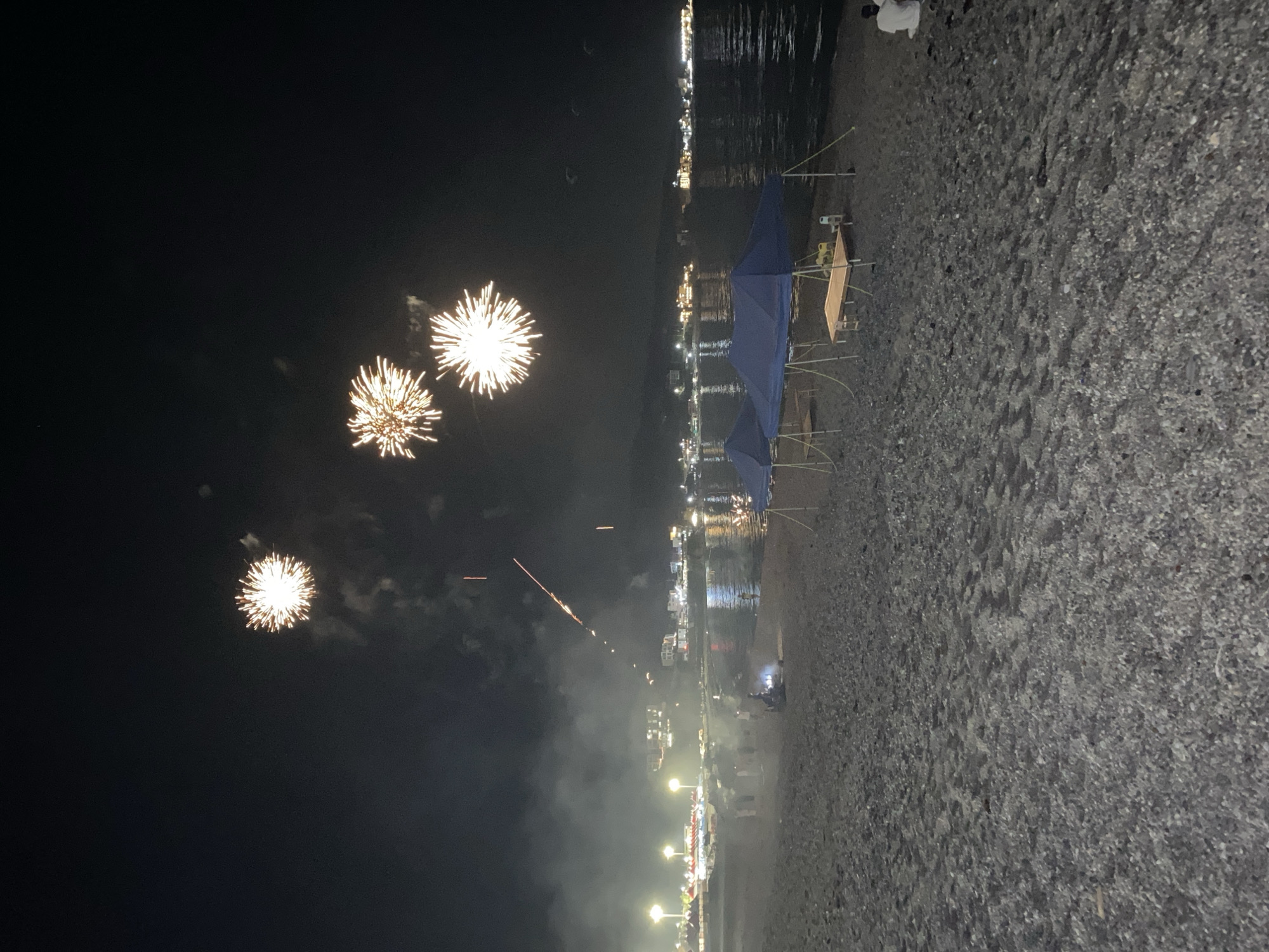 The image captures a nighttime scene on a beach in Gyeongju-si, South Korea, where fireworks are being set off over the ocean. In the foreground, a pebble beach is visible, with two blue tents set up on the sand. Tables and chairs are placed under the tents. A few people are seen sitting on the beach, one of whom is facing away from the camera. Further back, the coastline is lined with buildings and illuminated by lights. The sea reflects the lights from the shore and the fireworks. Several bright fireworks bursts are in the dark sky, with trails of smoke and light emanating from them. There are also smaller, less distinct streaks of light in the sky, possibly from other fireworks or fireworks being launched. The overall atmosphere is festive, indicating a celebration or event taking place on the beach.
