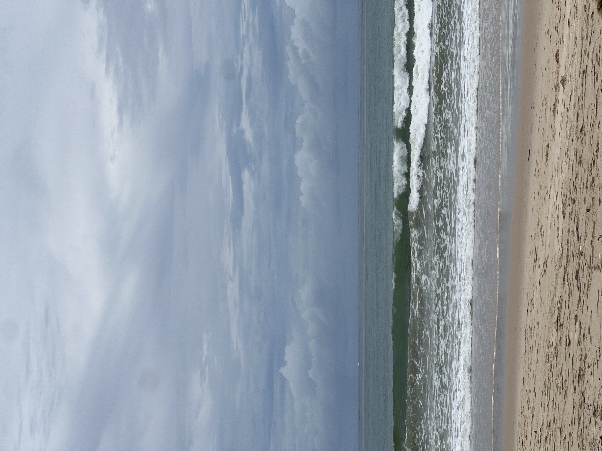 The image captures a beach scene. The sky is overcast with visible clouds. The sea displays waves breaking onto the shore. The sandy beach occupies the right portion of the image. The location is not specified. No people are visible.