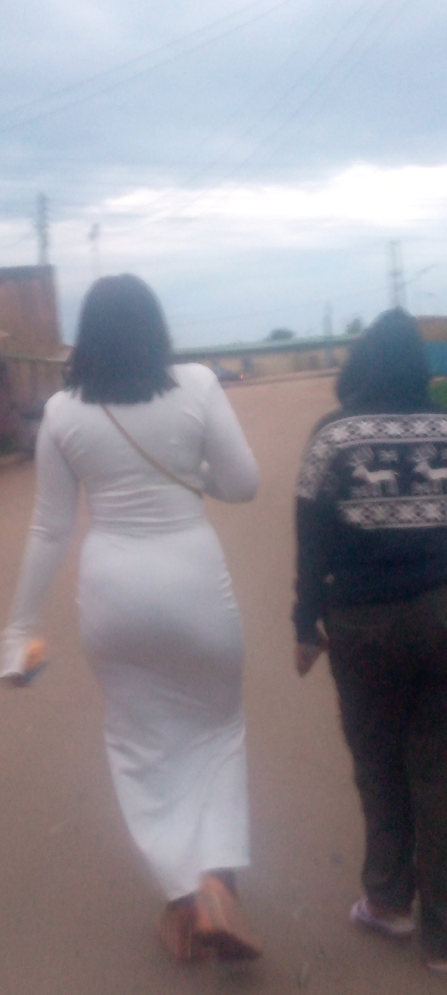 Two individuals, viewed from behind, walk on a paved surface under an overcast sky in an urban environment. The person on the left wears a long-sleeved white dress that hugs their body, with a thin strap of a bag visible across their shoulder. They are also carrying an orange object in their left hand. The person on the right wears a dark sweater with a white patterned design featuring deer and snowflakes, and dark pants. Their feet are shod in light-colored shoes, one appearing purple. The background shows structures, power lines, and vegetation, suggesting an outdoor, possibly somewhat dilapidated, urban setting. The lighting is dim, indicative of cloudy weather.