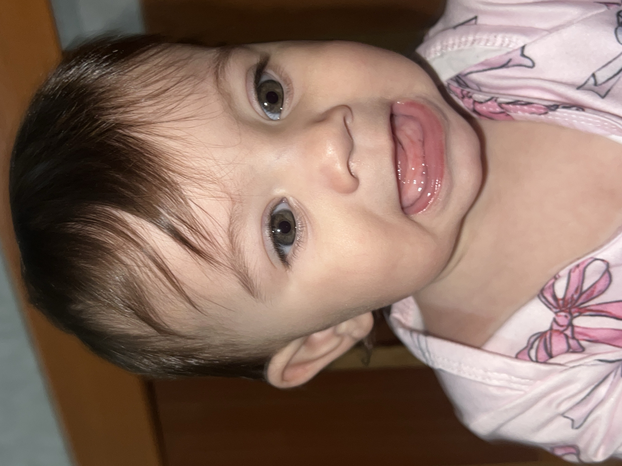 A close-up, angled portrait of a baby with dark brown hair and grey eyes. The baby is wearing a pink and white patterned onesie. The baby has their mouth open and tongue sticking out slightly. The background is blurred and appears to be a wooden surface. The location, Elena, Bulgaria, is not discernible from the image.