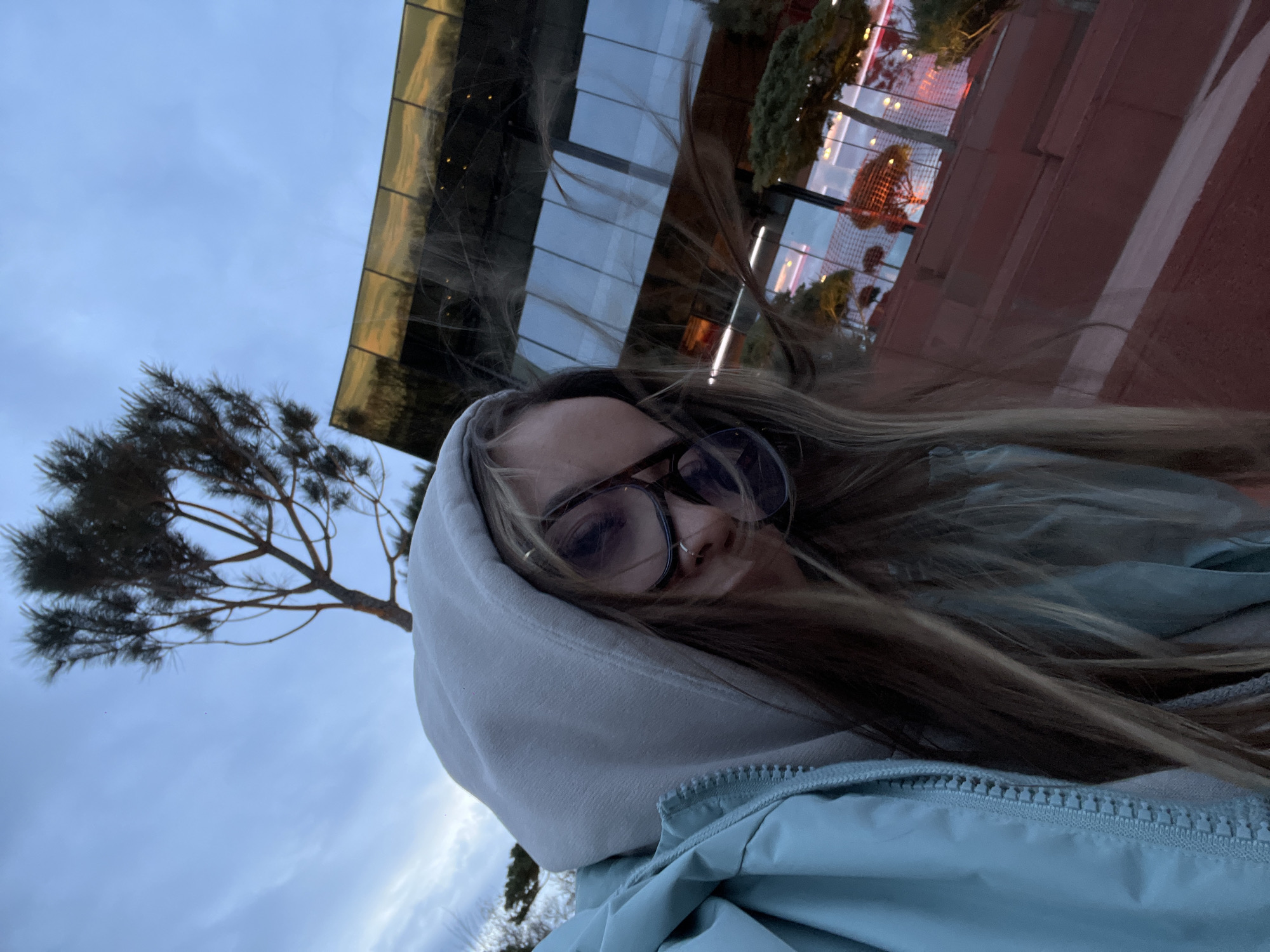 The image, rotated 90 degrees clockwise from its natural upright orientation, captures a casual selfie of a young person in an urban or semi-urban setting in Krasnodar, Russia. The perspective is from below, looking upwards.
The central subject is a person, likely a woman, wearing dark-rimmed round glasses and a light-colored (possibly light grey or beige) hooded sweatshirt with the hood up. Over the hoodie, she wears a light blue or teal zip-up jacket. Her long, light brown hair appears wind-swept, flowing around her face. A subtle nose piercing is visible. She looks directly at the camera with a neutral or slightly contemplative expression.
In the background, a partially cloudy sky in shades of light blue and white dominates the upper left. To the left, a tree with dark, sparse, pine-like foliage is silhouetted against the sky. To the right, a modern building with dark, reflective glass or metal panels is visible, catching warm, possibly golden or reddish, reflections. Below the building, some red and orange elements, possibly festive lights or architectural features, add a touch of color. The lighting suggests either an overcast day or the "blue hour" of late afternoon or early evening.
No specific event is taking place other than the act of taking a selfie. The image conveys a sense of being outdoors on a possibly breezy day. There is no visible text in the image.