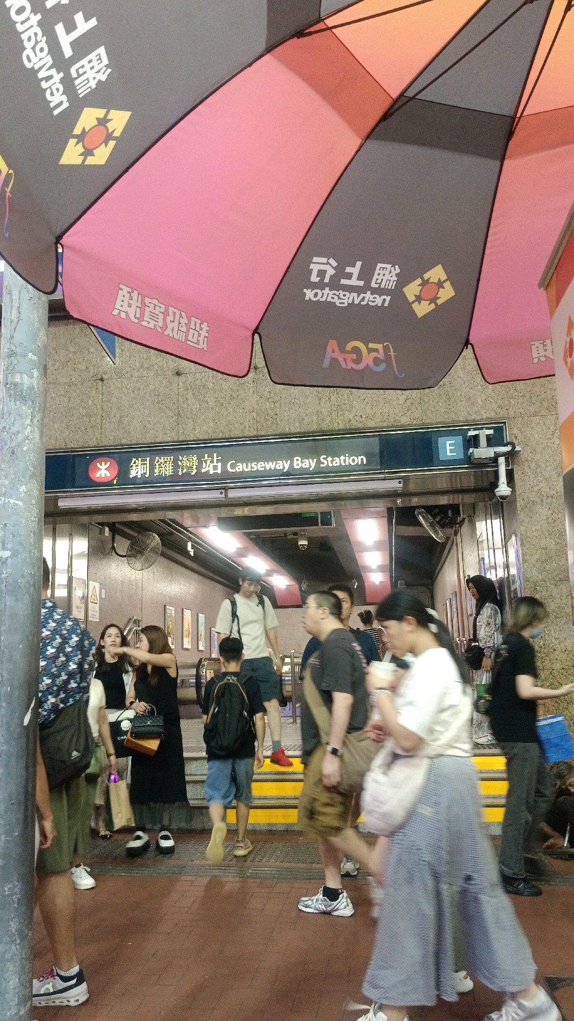 This image depicts the entrance to Causeway Bay Station in Hong Kong, China. The scene is crowded with people, suggesting a busy urban environment. Large, colorful umbrellas are suspended overhead, likely for shade or decoration. The station entrance is clearly marked with signage in both Chinese and English. People are seen walking in various directions, some carrying bags, indicating movement and activity. The ground is paved with reddish tiles. A metal pole is visible in the foreground, and the walls of the station are made of stone. The overall impression is one of a bustling public transportation hub.