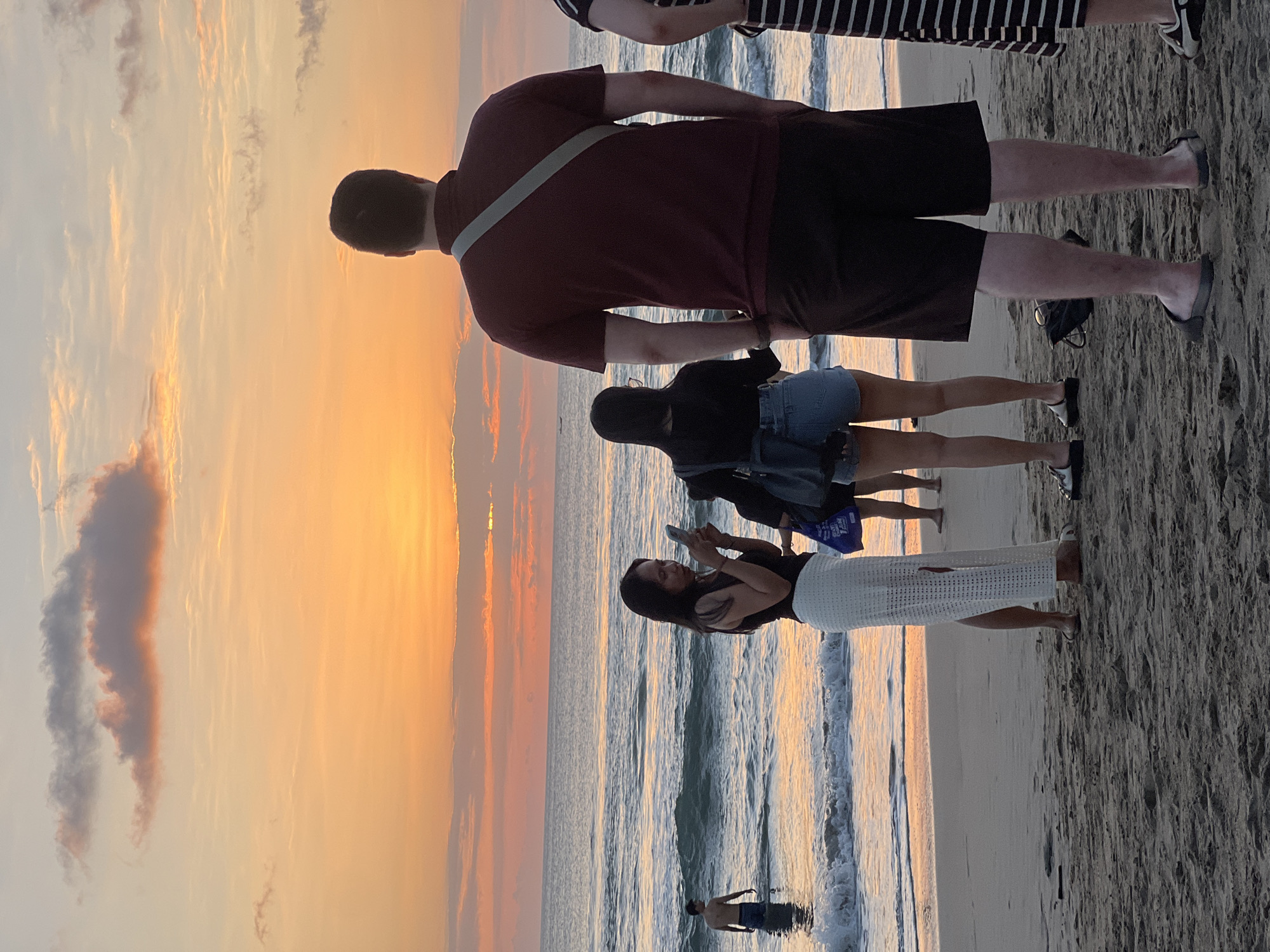 A group of people stand on a sandy beach in Kuta, Indonesia, facing the ocean as the sun sets. The sky is ablaze with shades of orange and yellow, with scattered clouds tinged with pink. Waves gently roll onto the shore. In the foreground, a man in a maroon polo shirt and dark shorts stands with his back to the camera, a light-colored strap across his shoulder. To his left, a woman in a white skirt and dark top holds up a phone, seemingly taking a picture. Beside her, another woman in denim shorts and a dark top also holds a phone. Further out in the water, a man with a bare chest stands waist-deep. To the right of the man in the maroon shirt, the striped dress of another person is visible. The sand is textured, with footprints and small debris. The overall atmosphere is one of calm and observation of the natural spectacle.