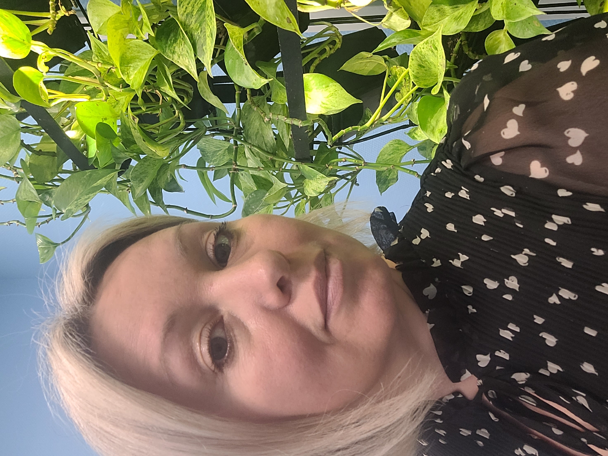 A woman with blonde hair and fair skin is the primary subject, looking directly at the camera from a low angle. She is wearing a black blouse with white heart-shaped patterns. Above her, a green plant with variegated leaves hangs from a black metal structure, appearing against a light blue background, which may be the sky. The location is not discernable from the image, and there is no information to suggest it is in Izhevsk, Russia. There is no visible action or interaction occurring in the image.