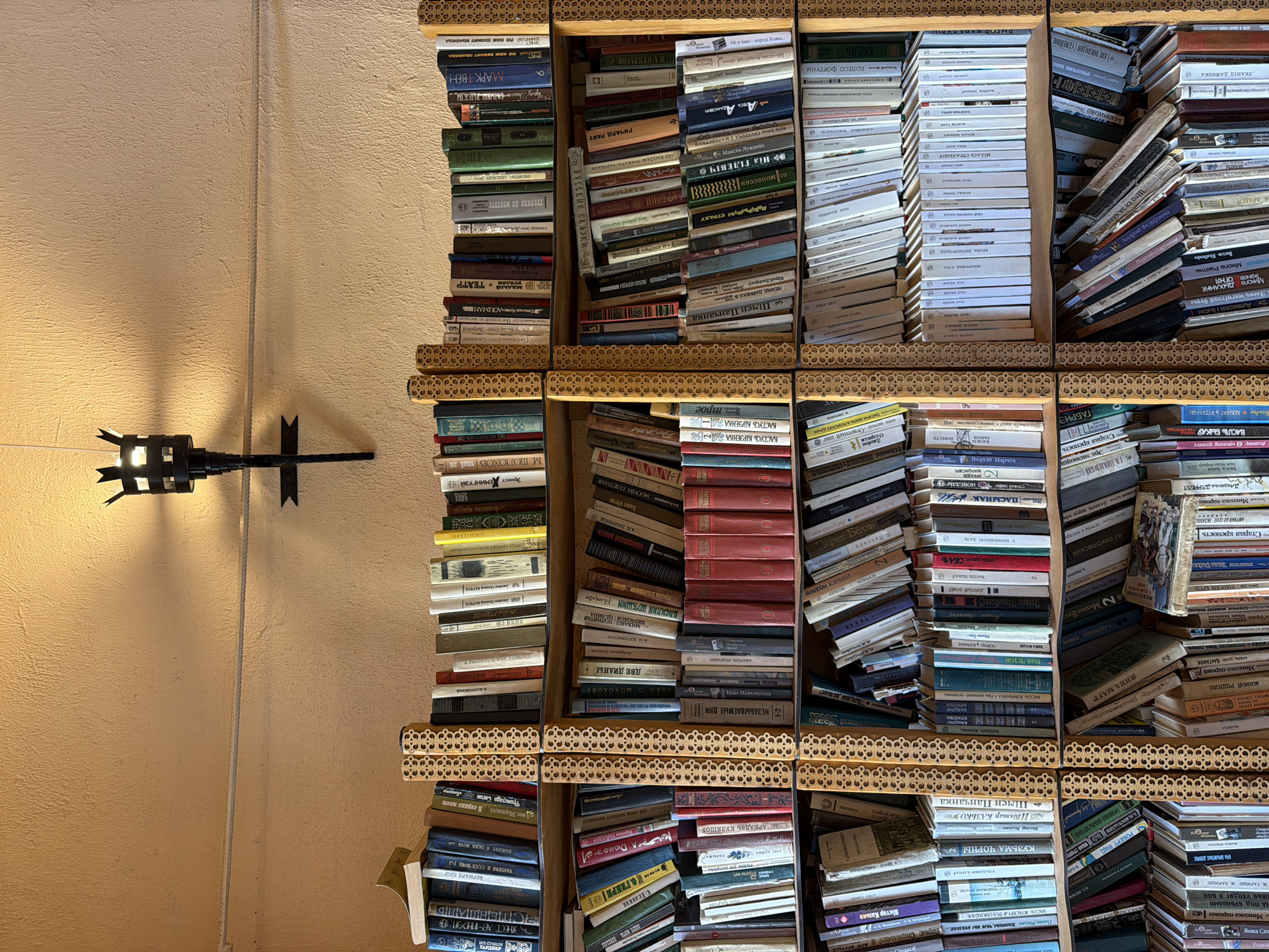 The image depicts a wall with a textured, light orange finish. Mounted on the wall, approximately two-thirds of the way up, is a black, wrought-iron sconce holding a torch-like light fixture with a flame visible. Below the sconce, the wall is lined with several wooden bookshelves, filled with numerous books. The bookshelves are divided into sections by decorative, golden wooden dividers with a floral pattern.
The books are arranged in rows, with their spines facing outwards. The books vary in size, color, and thickness. Some shelves are densely packed, while others have fewer books. The lighting suggests it is either daytime or the sconce is the primary light source.
Specific books visible include titles in Cyrillic script, indicating a likely location in a Slavic-speaking country. The presence of a wall sconce and extensive bookshelves suggests an interior setting such as a library, study, or a room in a house. The overall impression is one of a well-stocked and perhaps slightly dated library or reading area.
The provided location, Літвенскі сельскі Савет, Belarus, is a rural administrative unit. This context suggests the setting could be a rural library, a community center, or a private residence within this area. Without further information or visual cues, it is difficult to pinpoint the exact type of establishment.